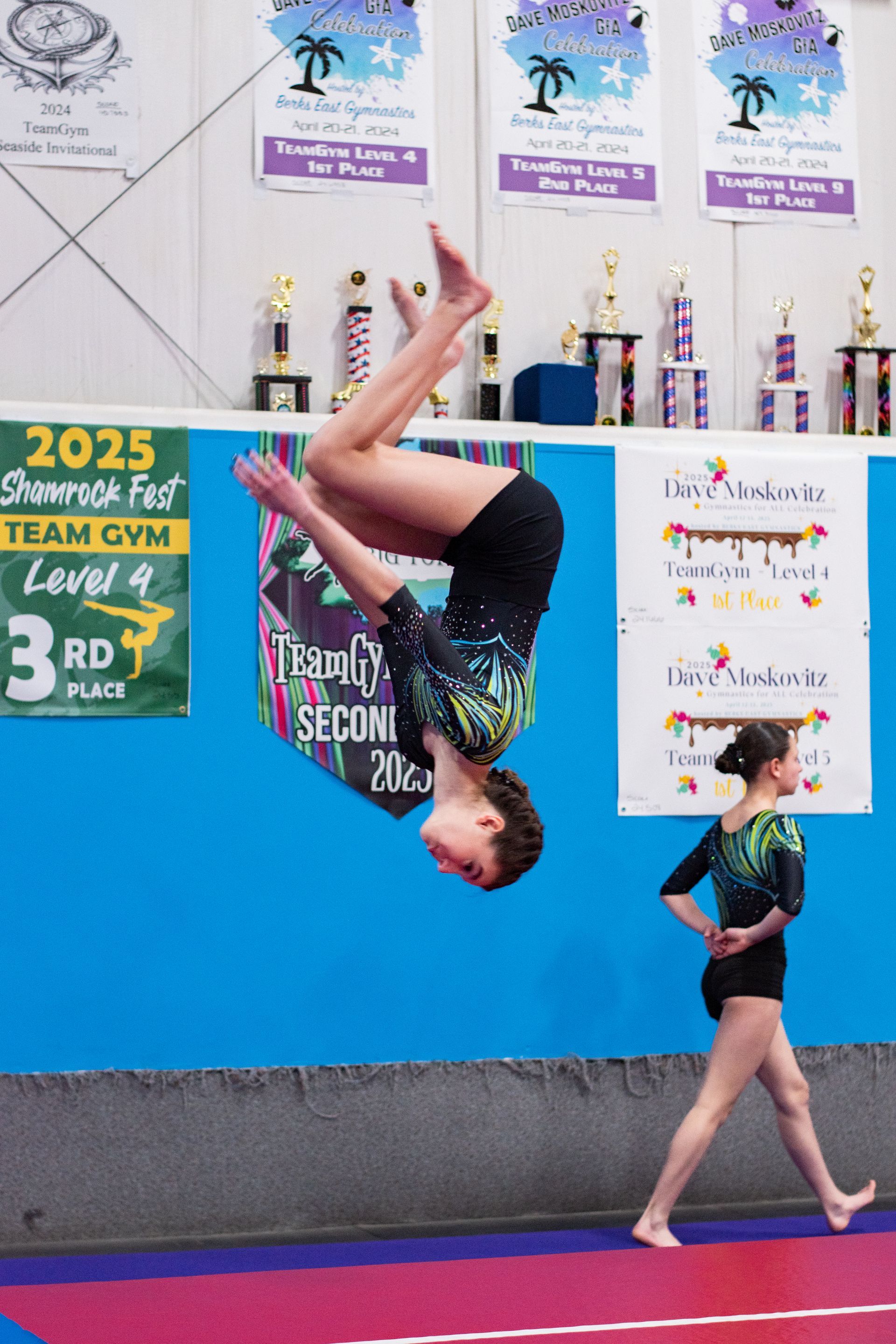 Gymnast mid-backflip on blue mat, with trophies and competition banners in the background.