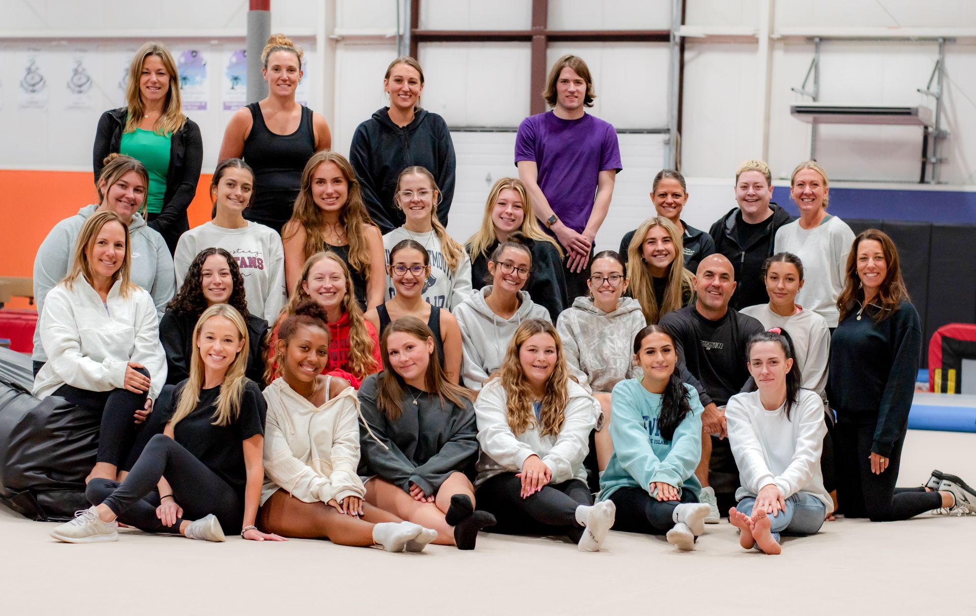 Group of people posing together in a gym, seated and standing in rows against a white wall.