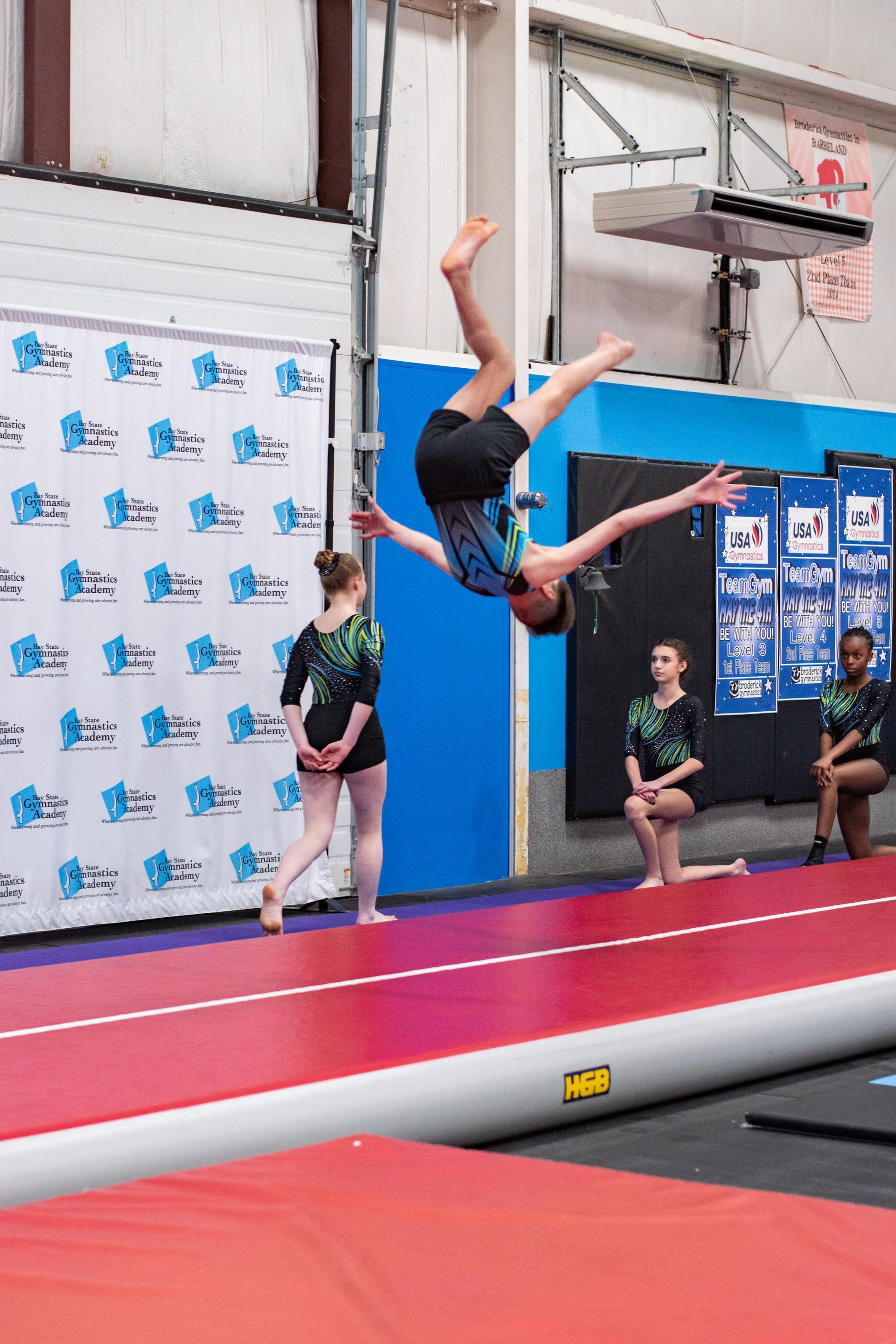 Gymnast mid-backflip over red mat in an indoor gym, with teammates watching nearby.
