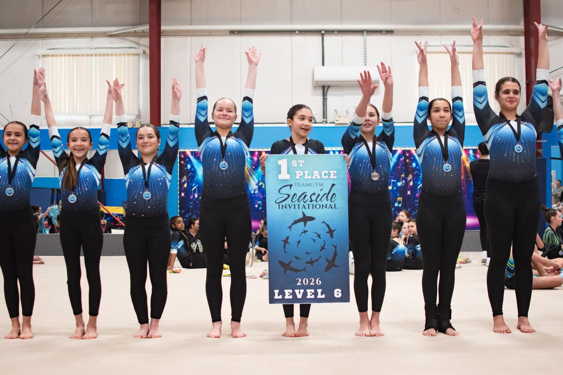 Gymnastics team in blue uniforms posing with raised arms, holding a “1st Place” sign on the floor.