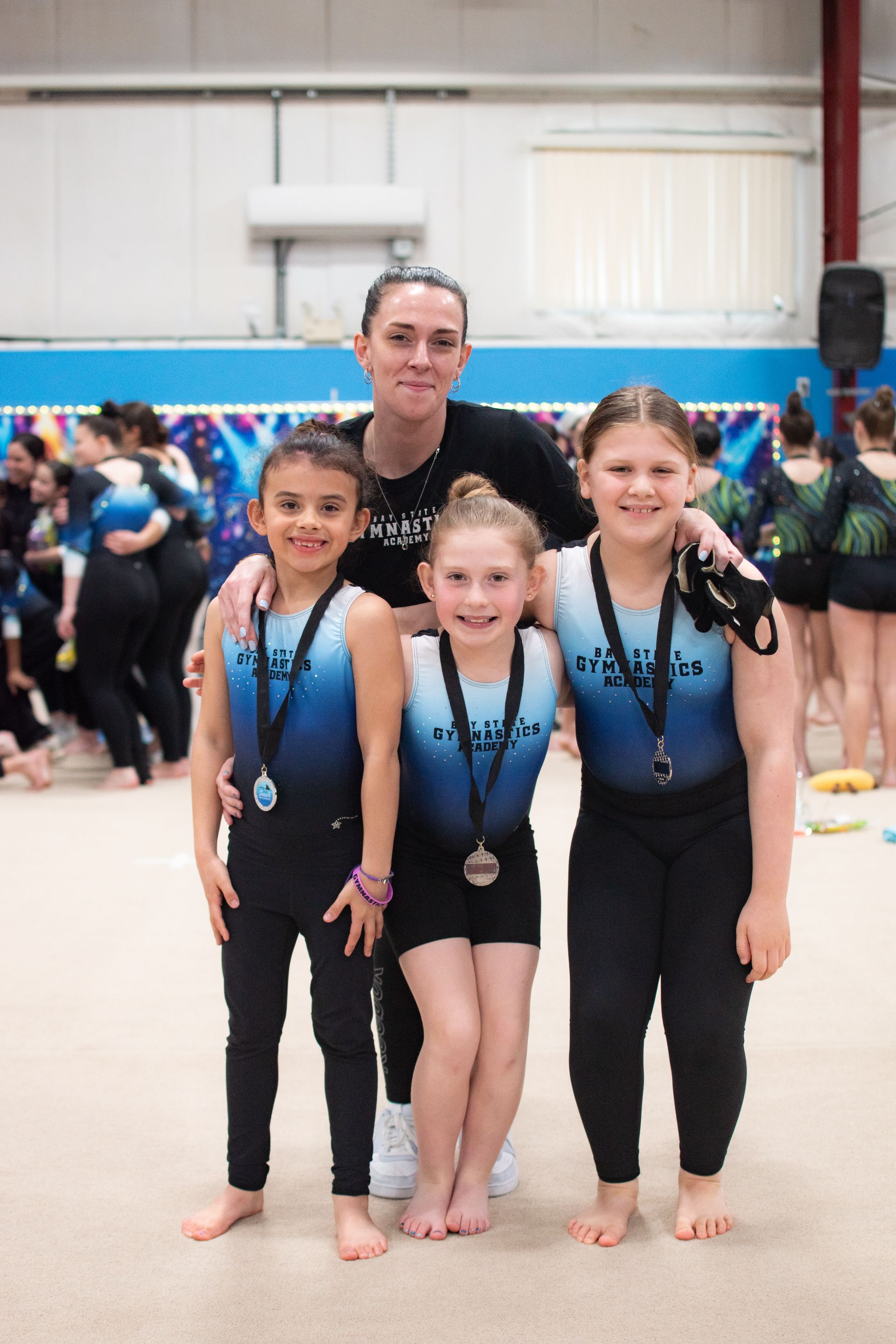 Four young gymnasts with medals pose in matching blue-and-black leotards inside a gymnasium.