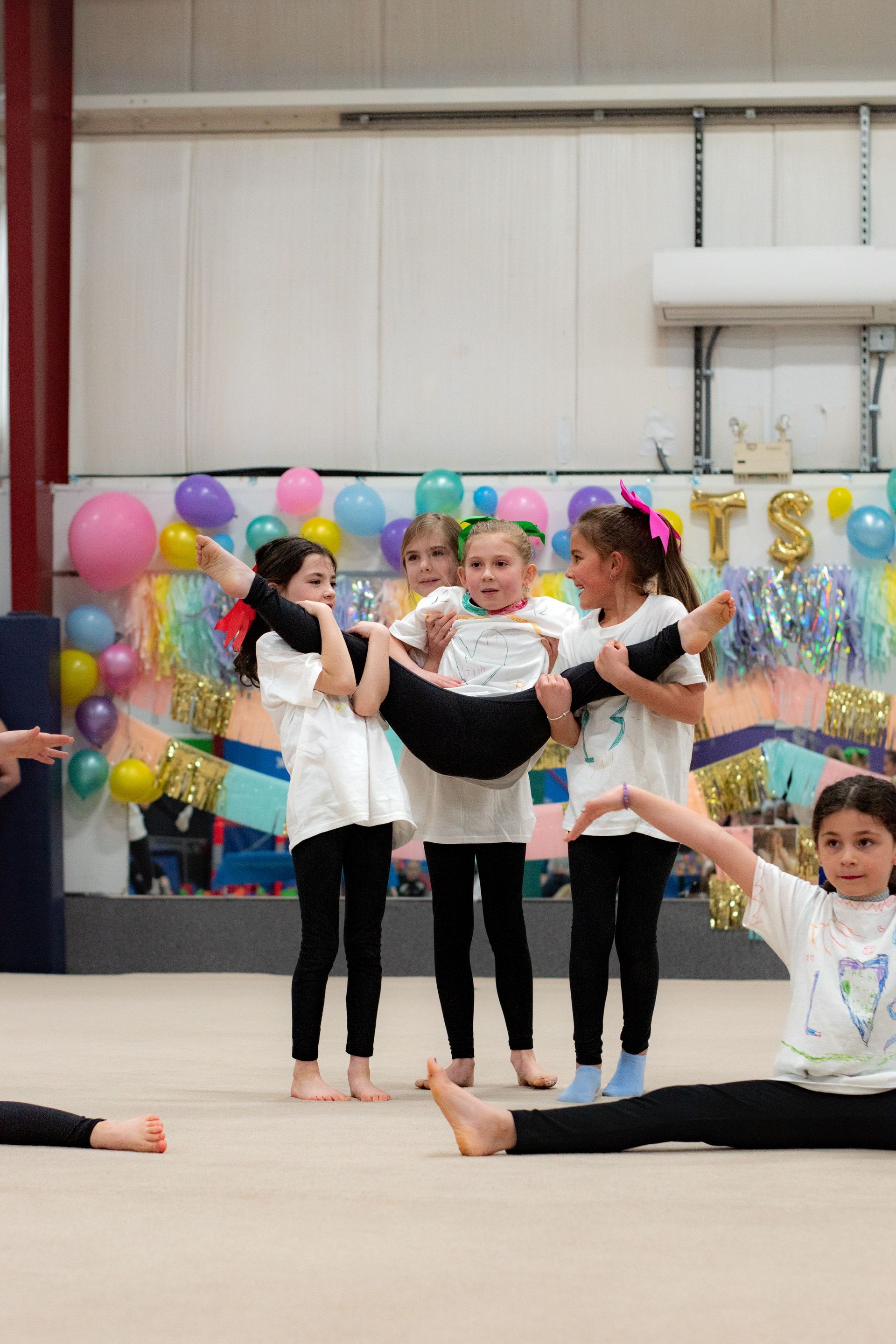 Children performing a group dance lift on a gym floor, with colorful balloons and a festive backdrop.