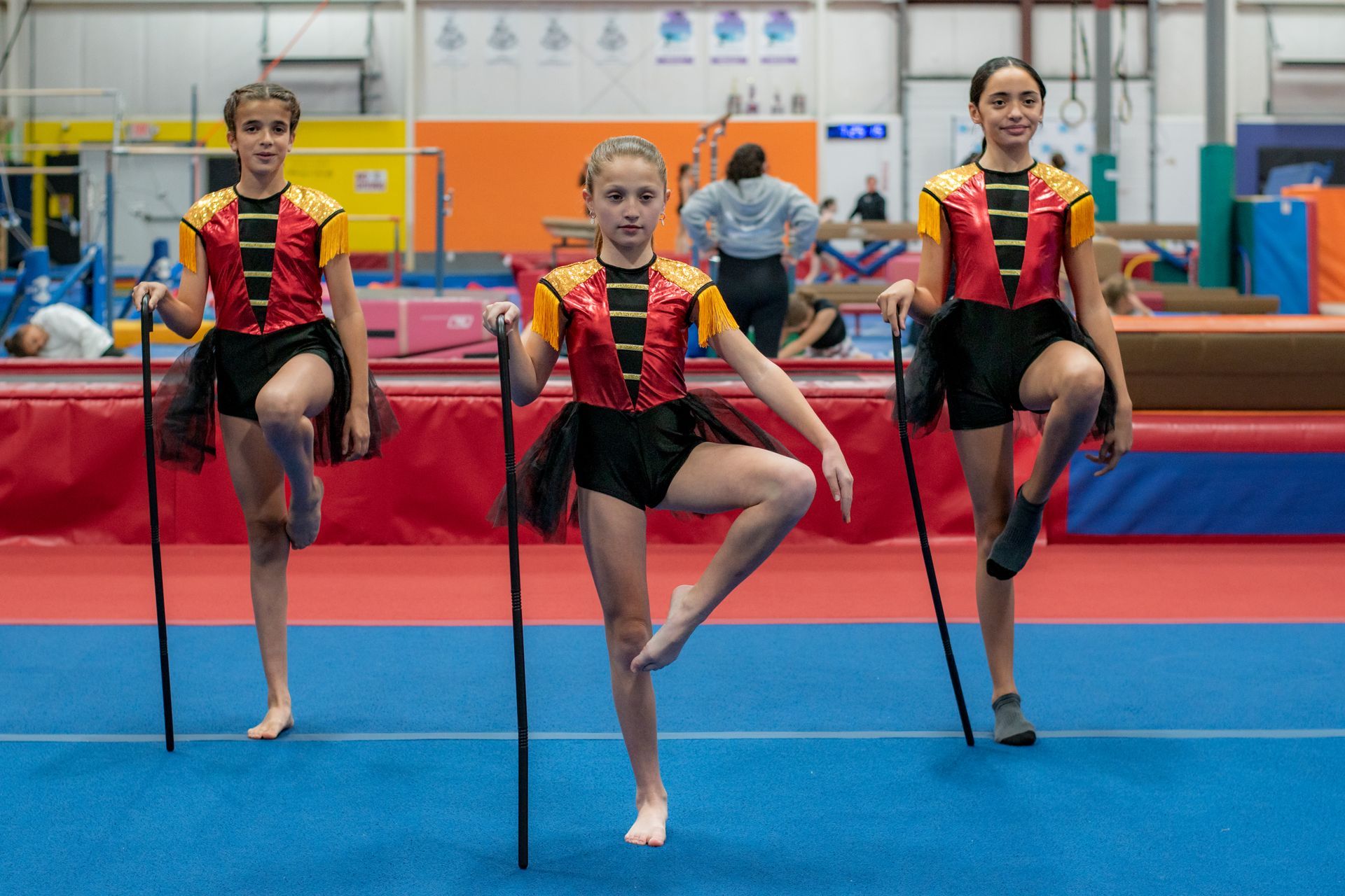 Three gymnasts in red and black leotards balance on canes in a gymnasium