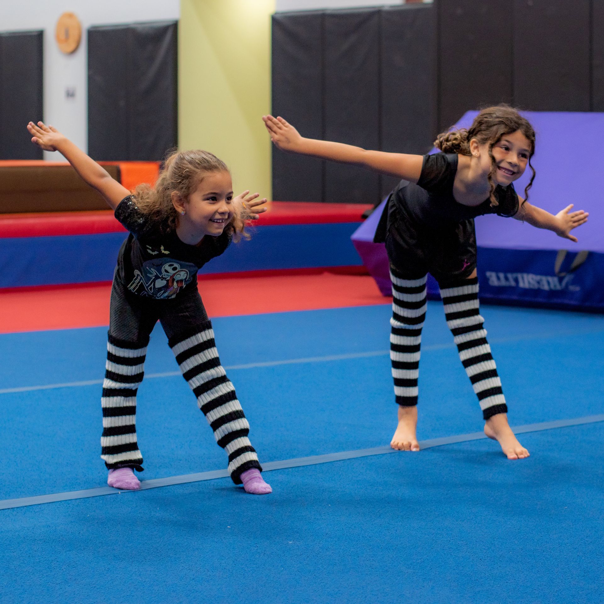 Two children balancing with arms outstretched on a blue gym mat in an indoor gym.