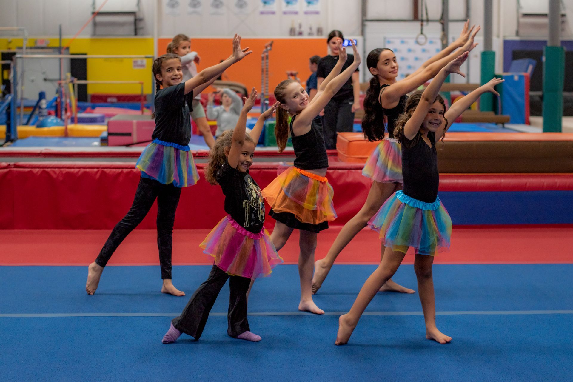 Five gymnasts in colorful skirts pose with arms raised on a blue floor in an indoor gymnasium.
