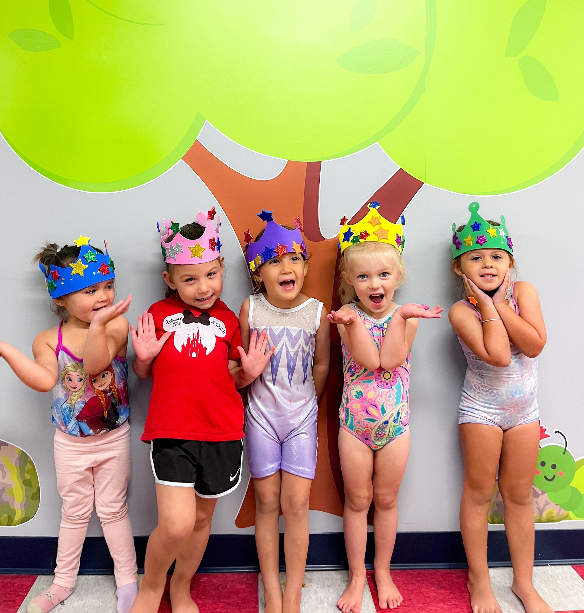 Five children in colorful party hats posing in swimsuits under a cartoon tree backdrop