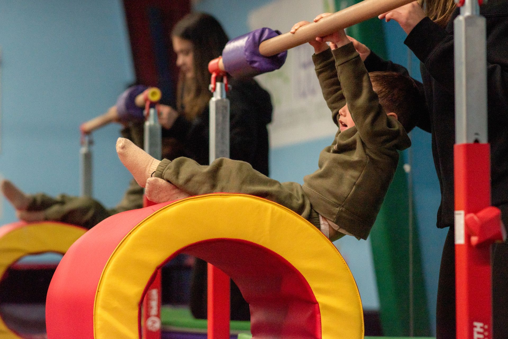 Children playing on colorful gym equipment, hanging upside down on bars in an indoor play area.