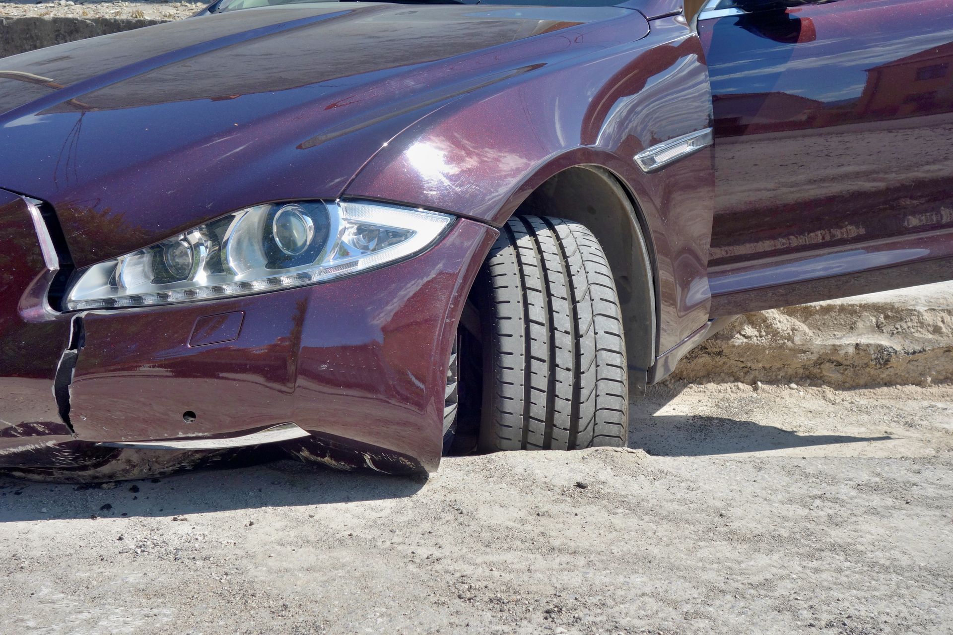 Front wheel of a car lodged in a deep pothole with visible damage to the bumper and tire. Front wheel of a car lodged in a deep pothole with visible damage to the bumper and tire.