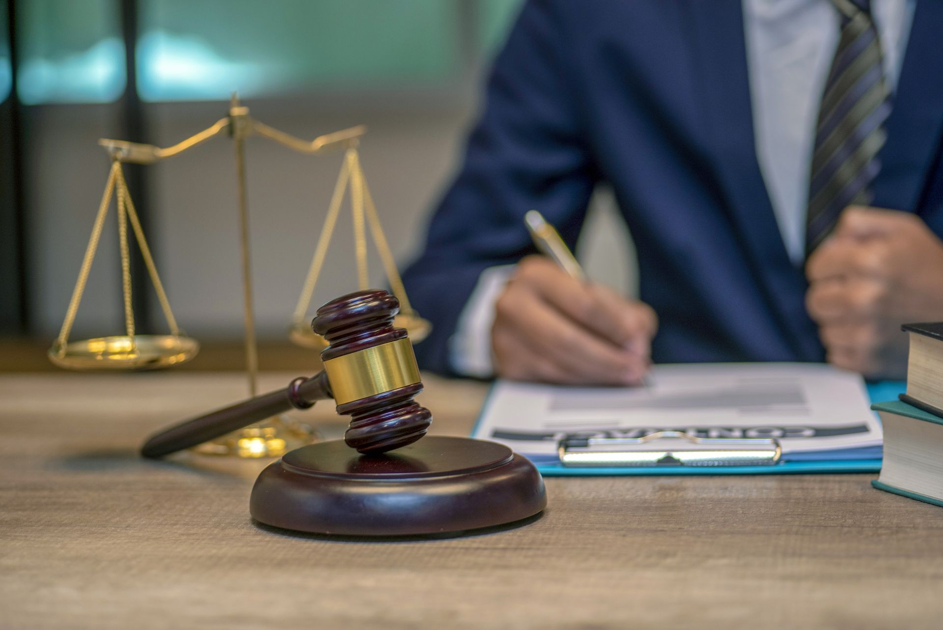 Gavel on desk in front of scales of justice and a person in a suit writing on a clipboard.