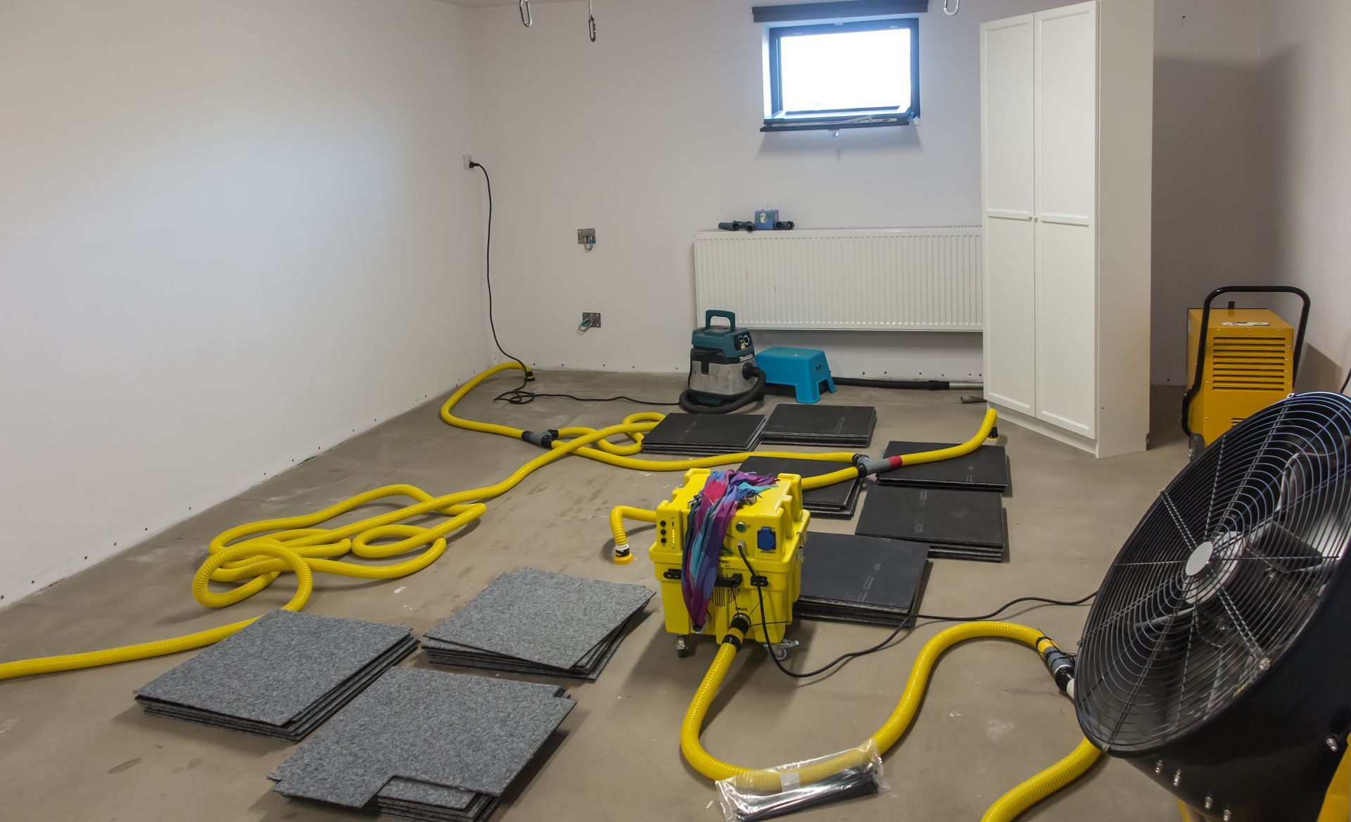 Basement room with construction tools, yellow hoses, floor panels, and a large fan near a window