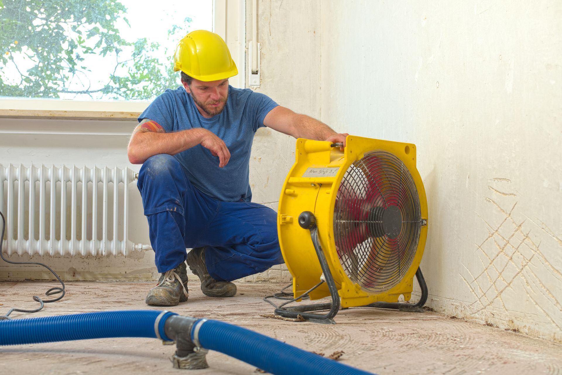 Worker in hard hat inspecting a yellow industrial fan beside blue pipes in a room under renovation