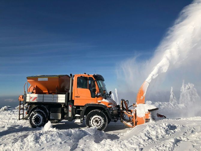 Orange snowplow clearing snow in a snowy landscape under a clear blue sky