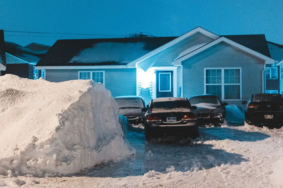 Snow-covered driveway with two parked cars in front of a lit suburban house at night