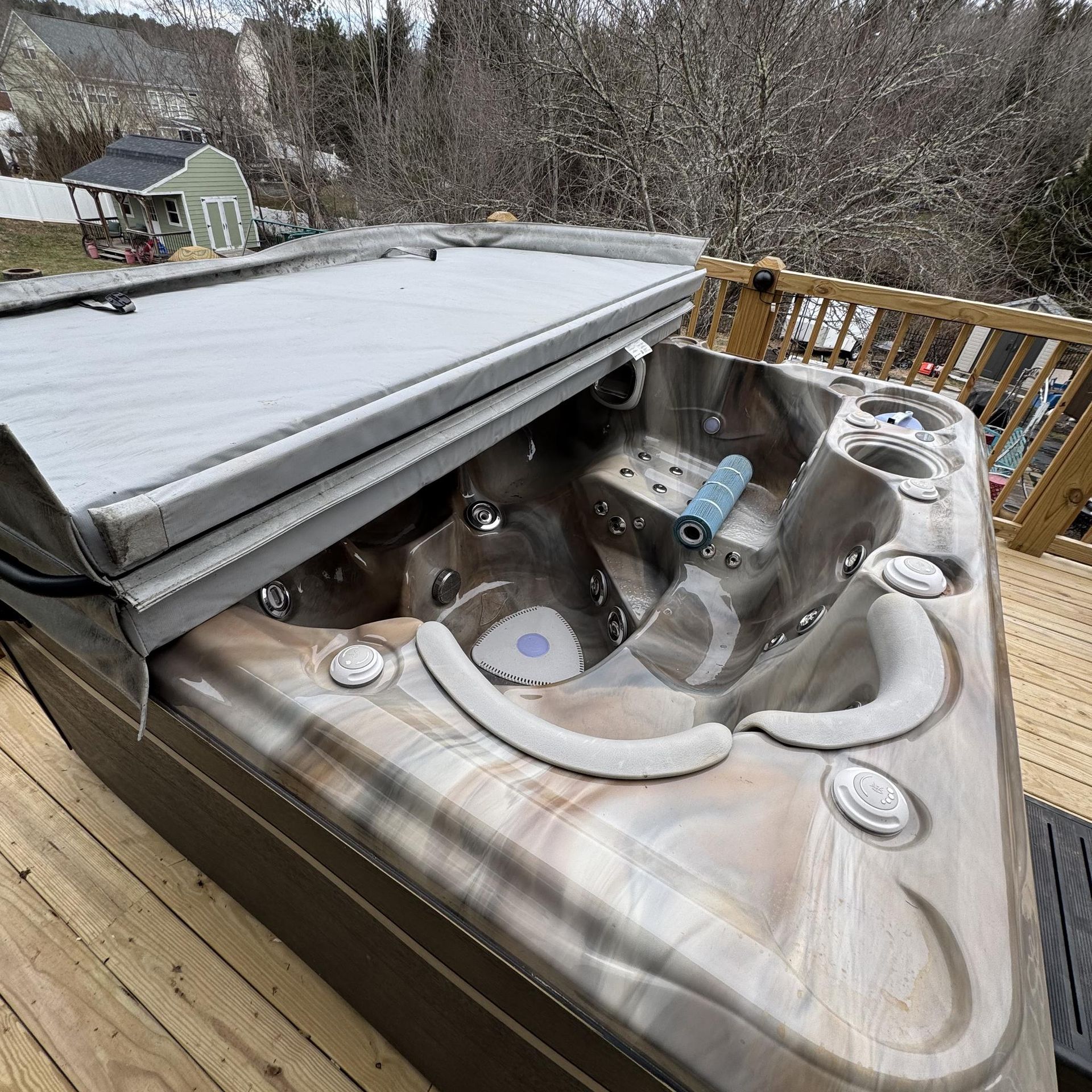 Hot tub with open lid on a wooden deck. The tub is brown with jets and controls. The cover is gray.