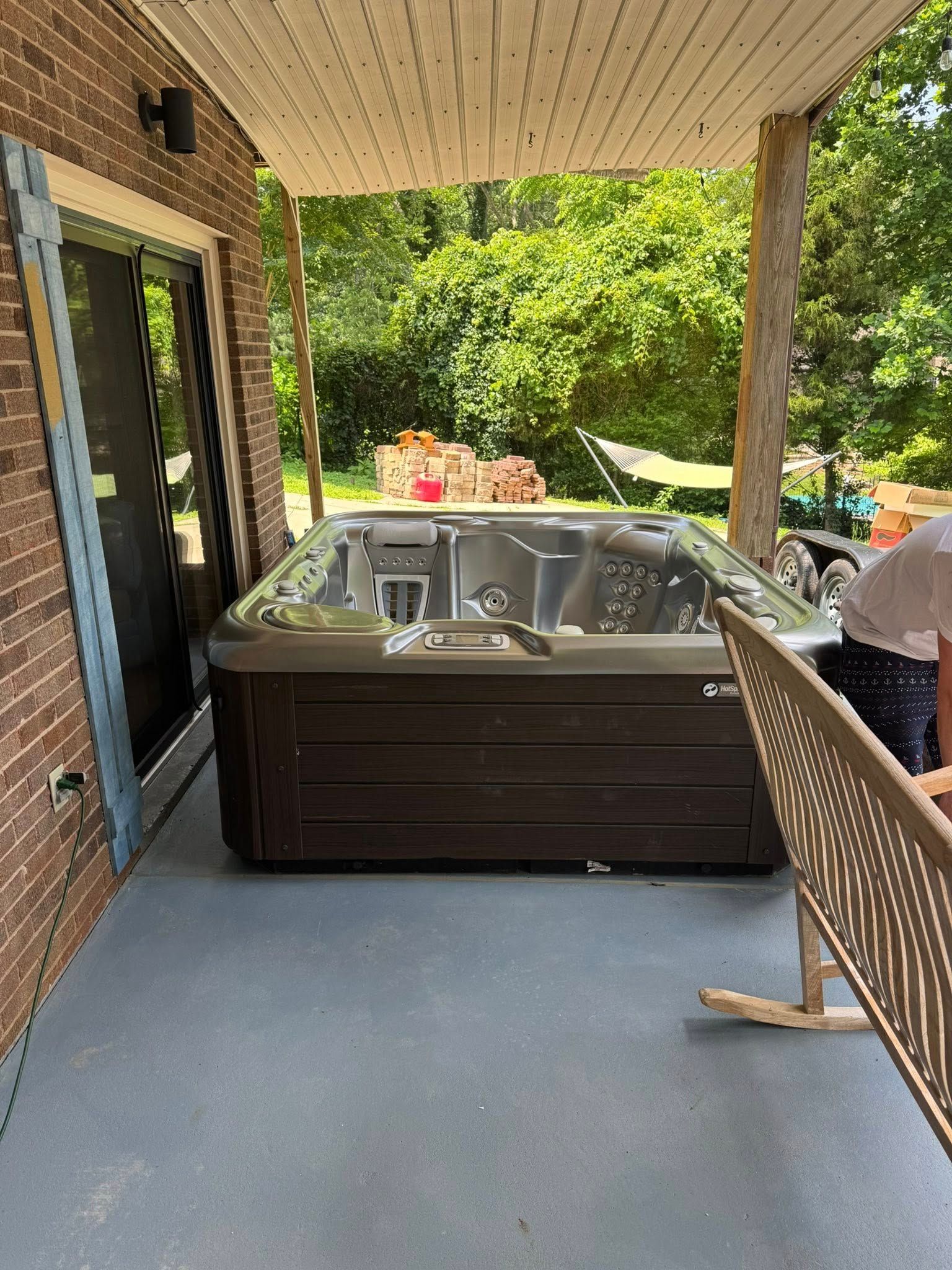 Hot tub on a porch under a roof. Brown exterior, gray floor, trees in the background.