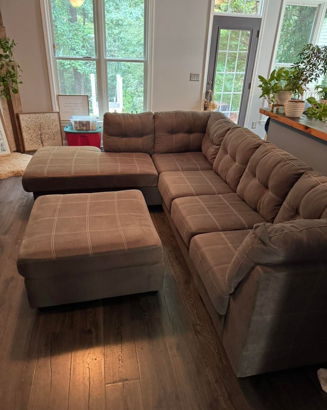 Brown sectional sofa and matching ottoman in a living room with a window and plants.