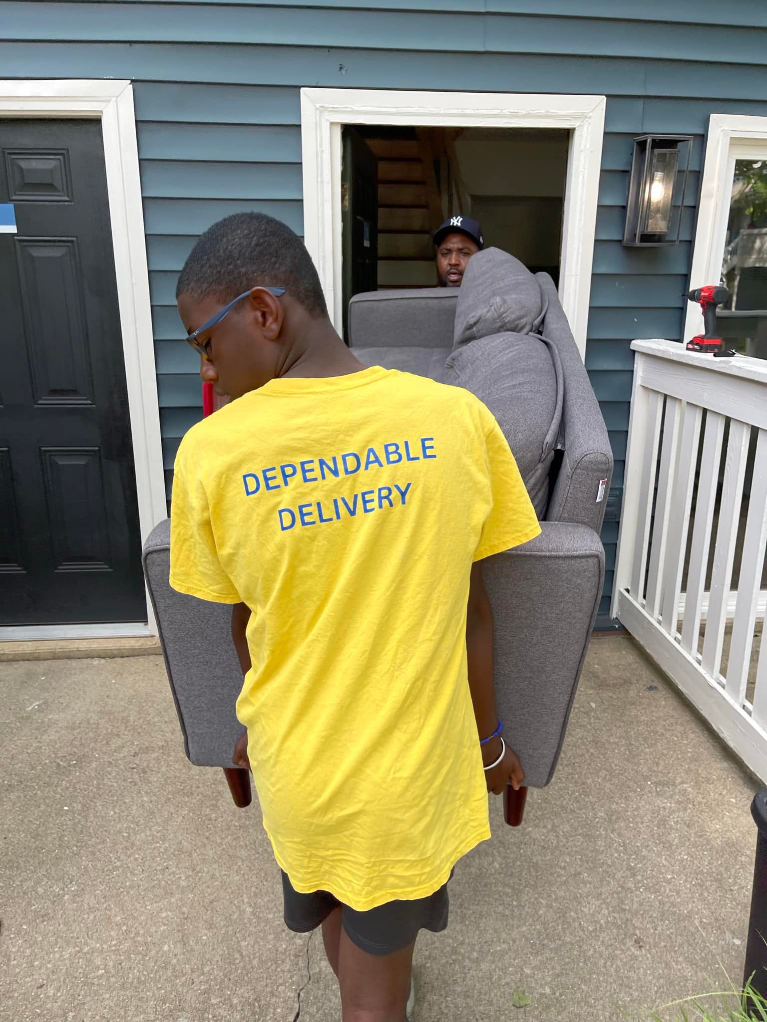 Person carrying a gray sofa out of a house doorway. The person wears a yellow 