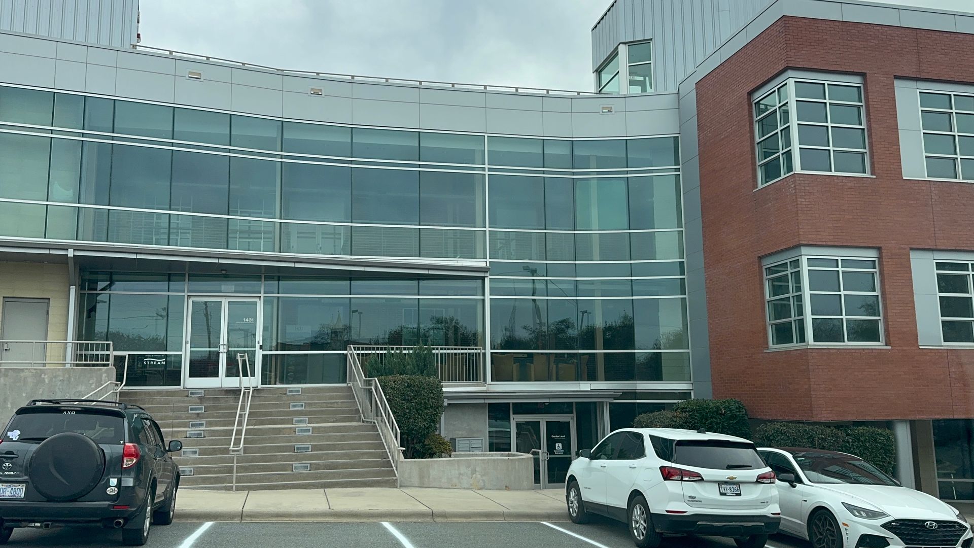 Modern glass and brick building with cars parked in front. Entrance stairs and windows visible.