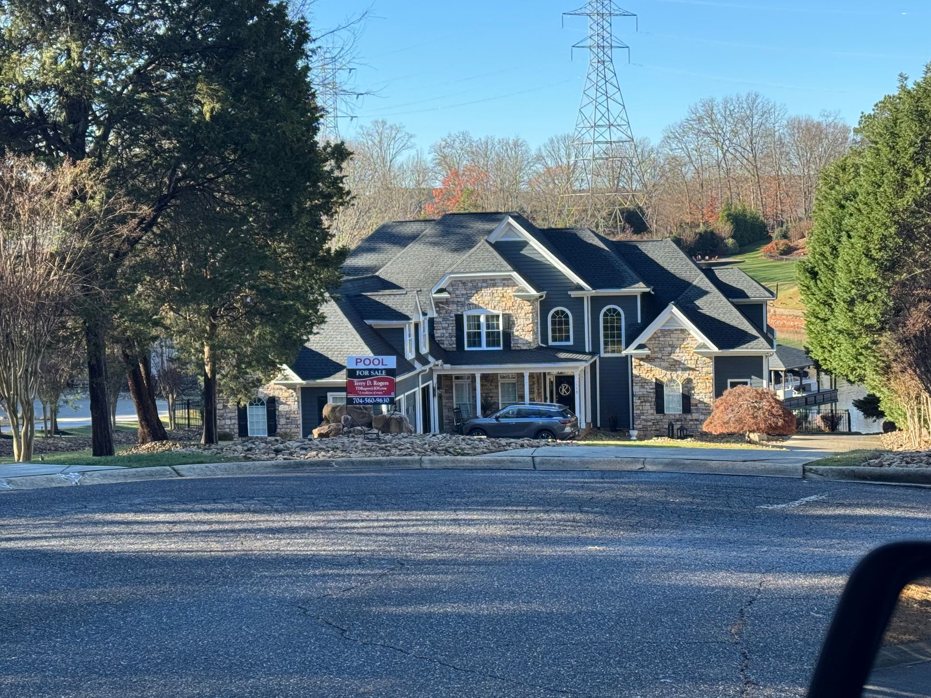 Two-story house with dark siding, stone accents, and a blue car parked in the driveway; power lines in the background.