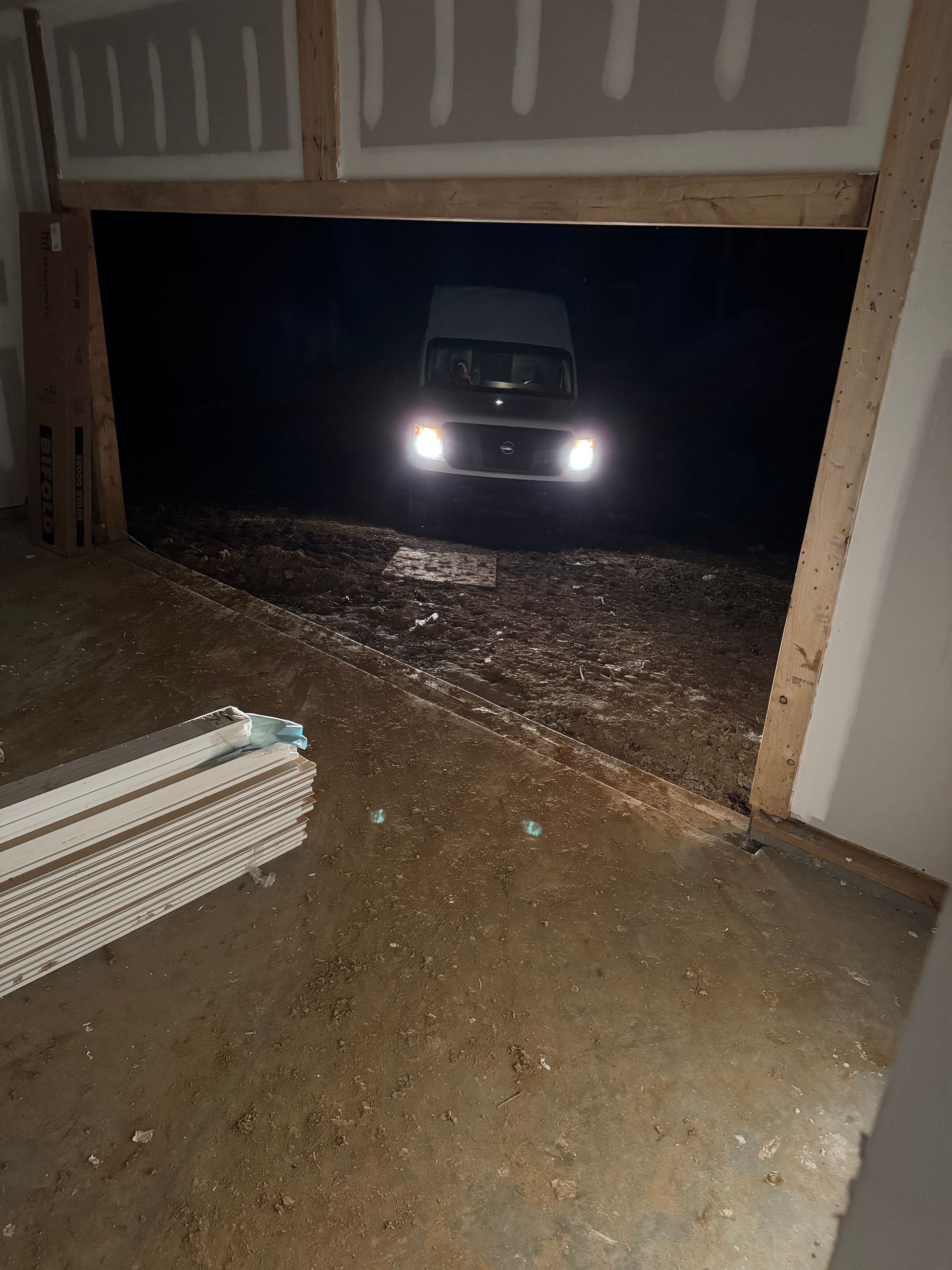 Van with headlights on, parked outside unfinished building. Dark setting, muddy ground.