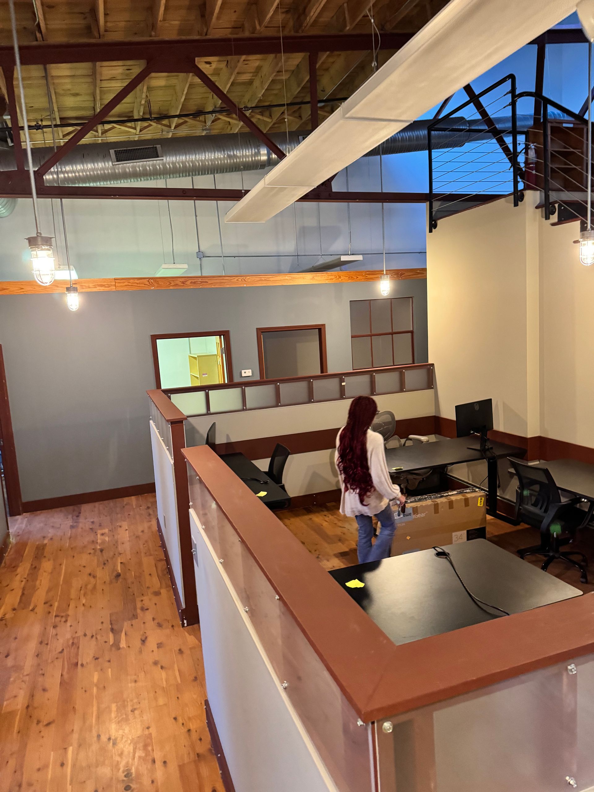 Office interior with a person walking past desks. Wooden floors, gray walls, and exposed beams.
