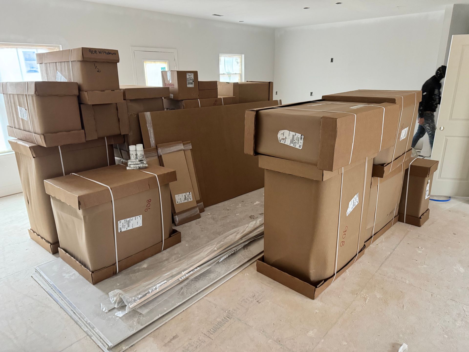 Boxes and building materials piled in an unfinished room; a person works in the background.