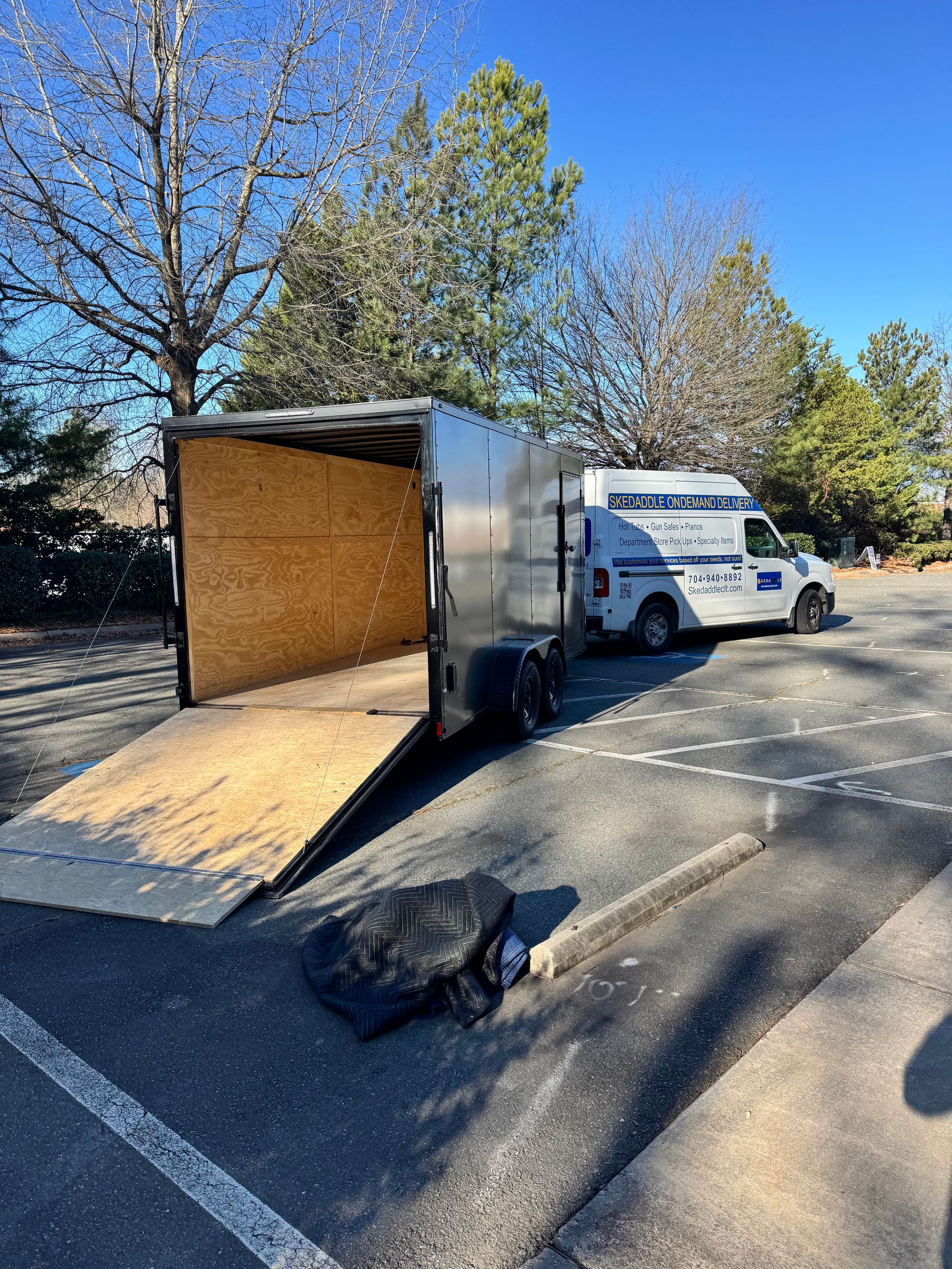Silver trailer with open ramp, parked next to a white truck in a paved lot. Blue sky.