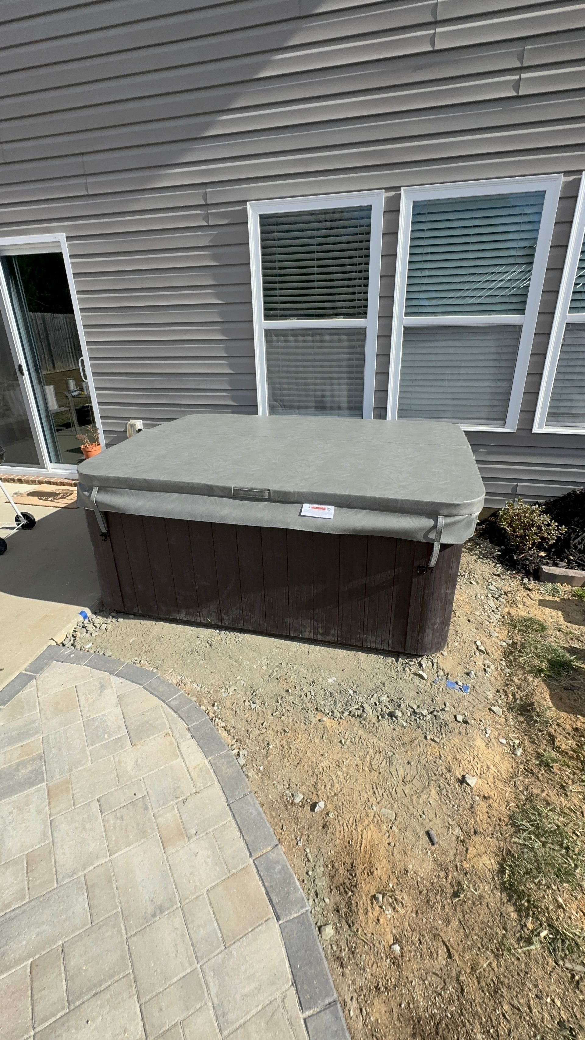 Hot tub with cover, brown siding, windows, and brick patio.