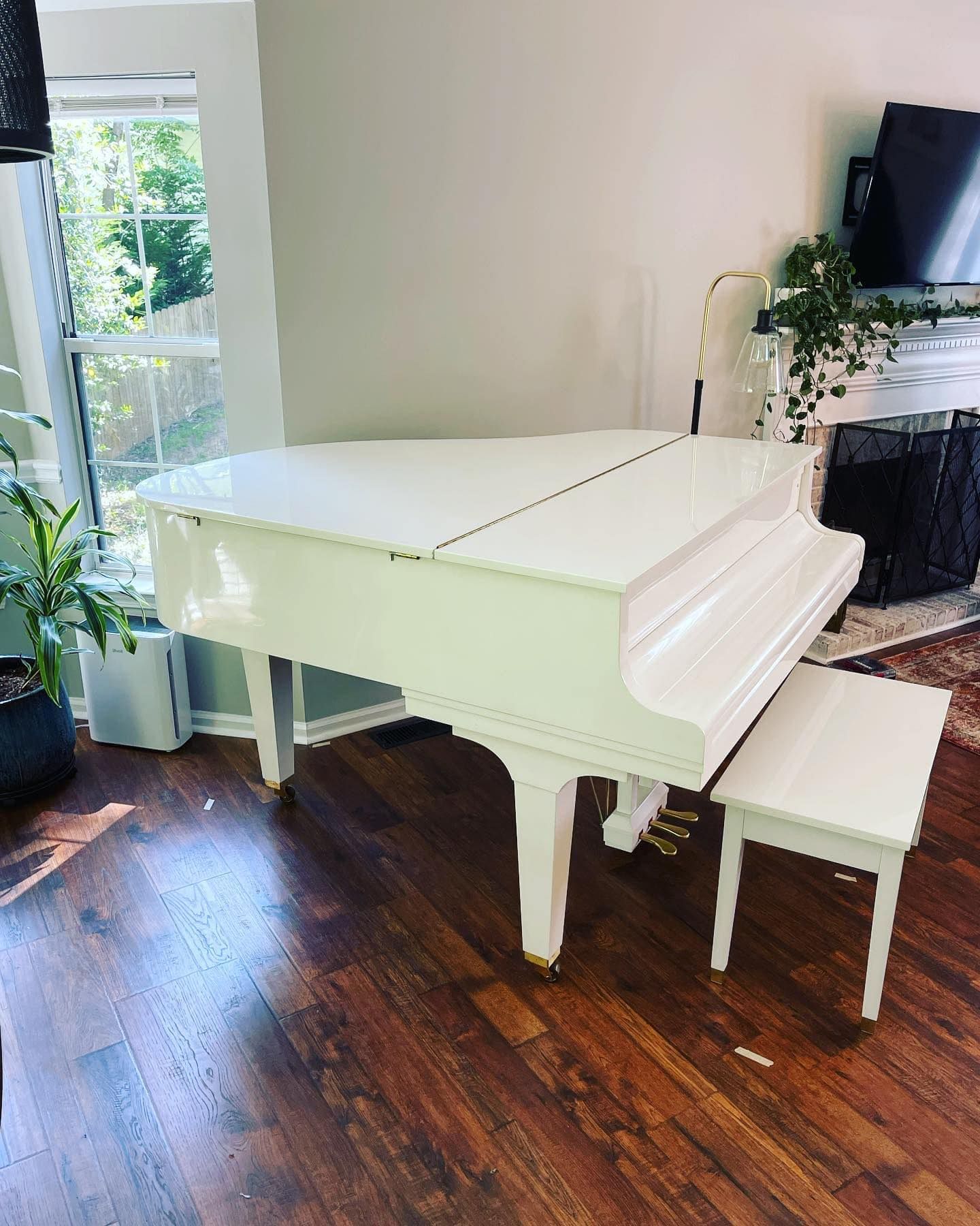 White grand piano and bench in a room with hardwood floors, a window, and a fireplace.