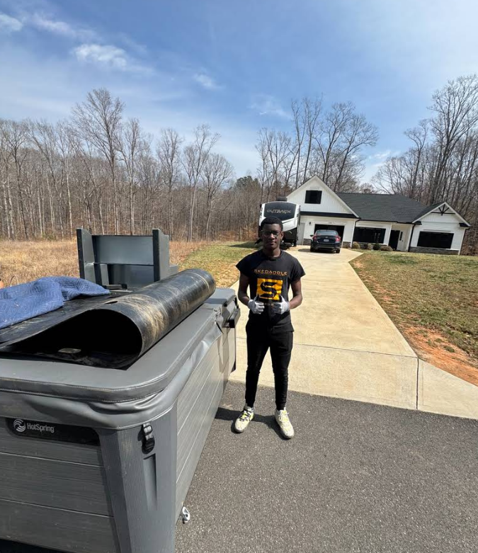 Person standing next to a hot tub in front of a house. Black car in the driveway. Outdoors on a sunny day.
