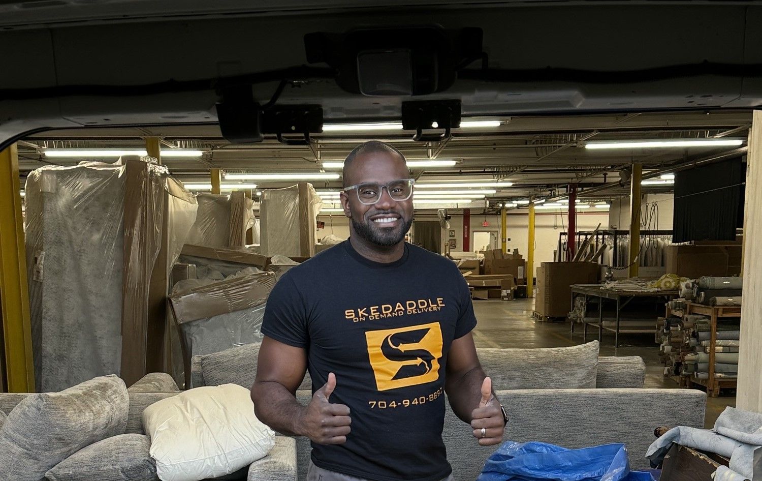 Man in black t-shirt giving thumbs up in a warehouse setting. Lots of materials and equipment visible.