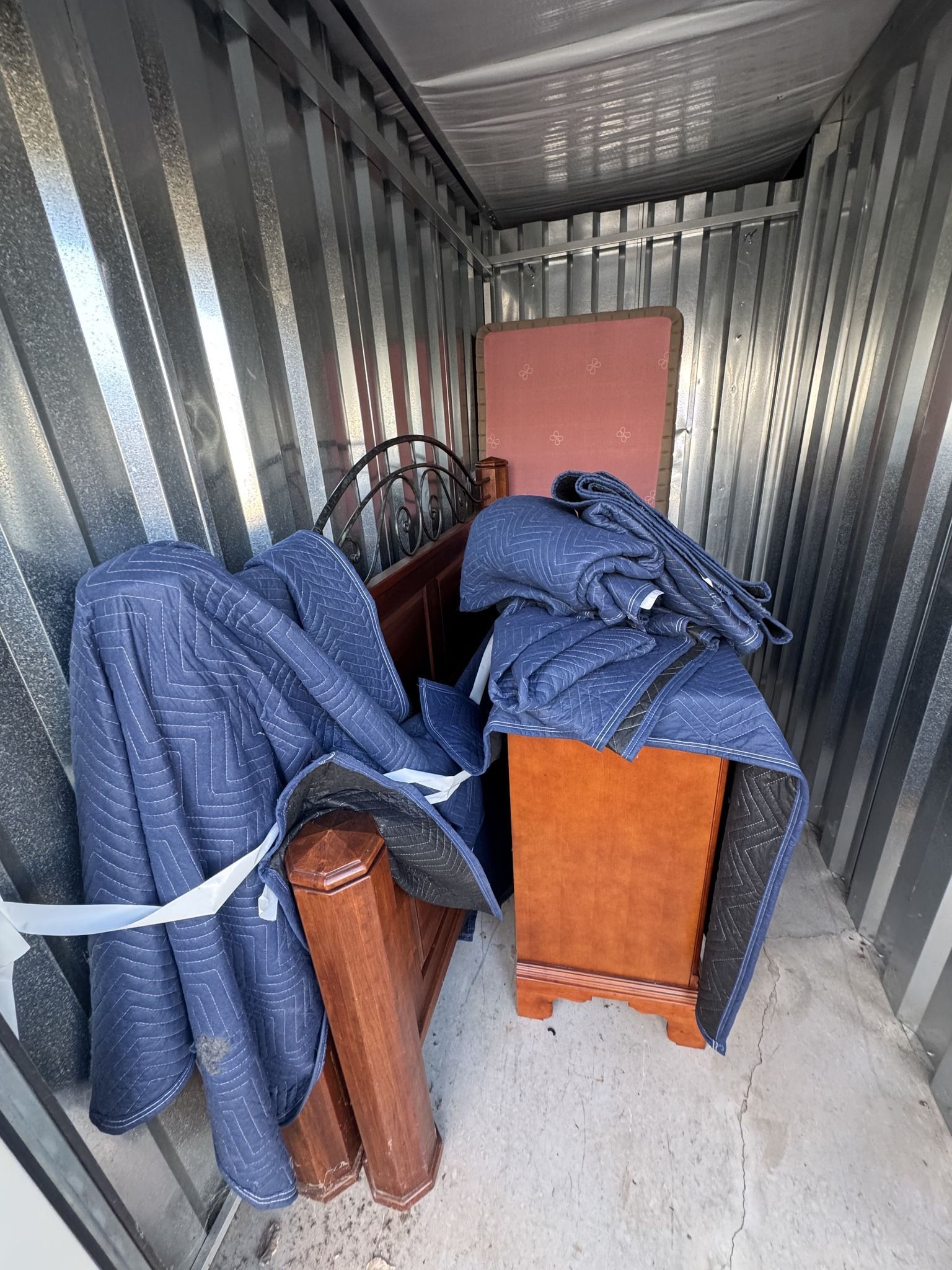 Storage unit interior with bed frame, dresser, and wrapped furniture.