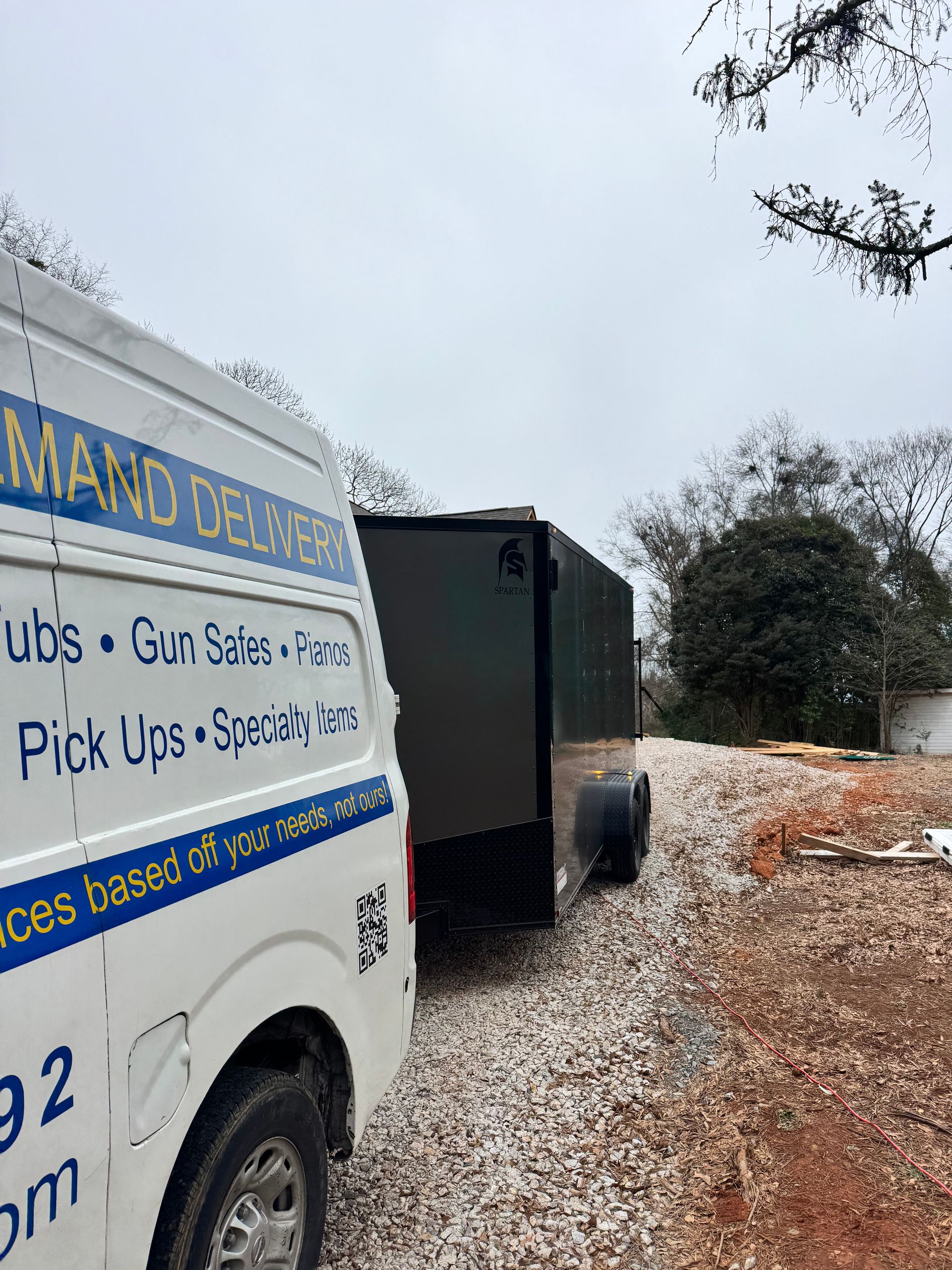 A white van with blue text is parked beside a dark trailer on a gravel driveway under an overcast sky.