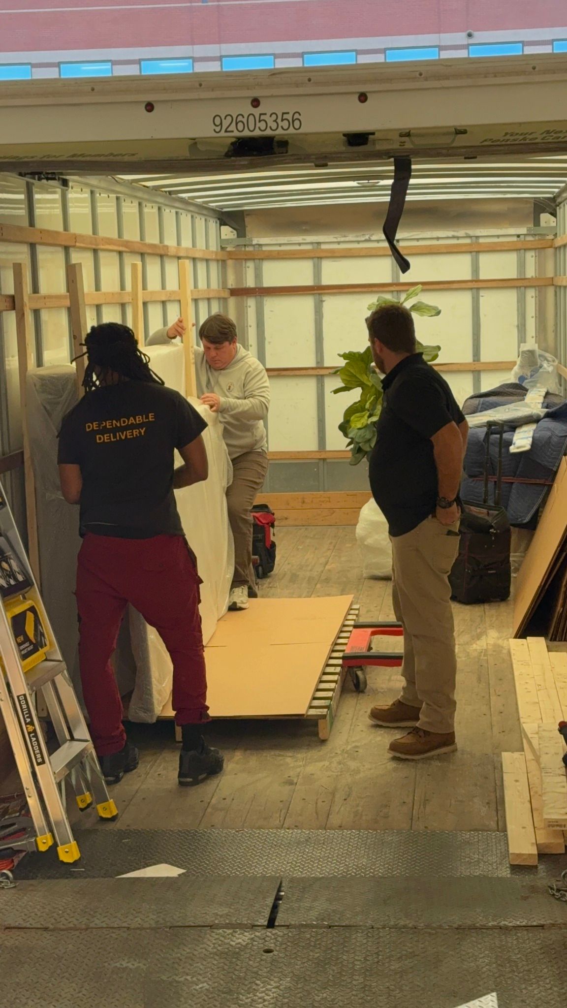 Three people loading furniture into a moving truck. One man in a black shirt, khaki pants, brown boots watches. Two others work with large white boxes.