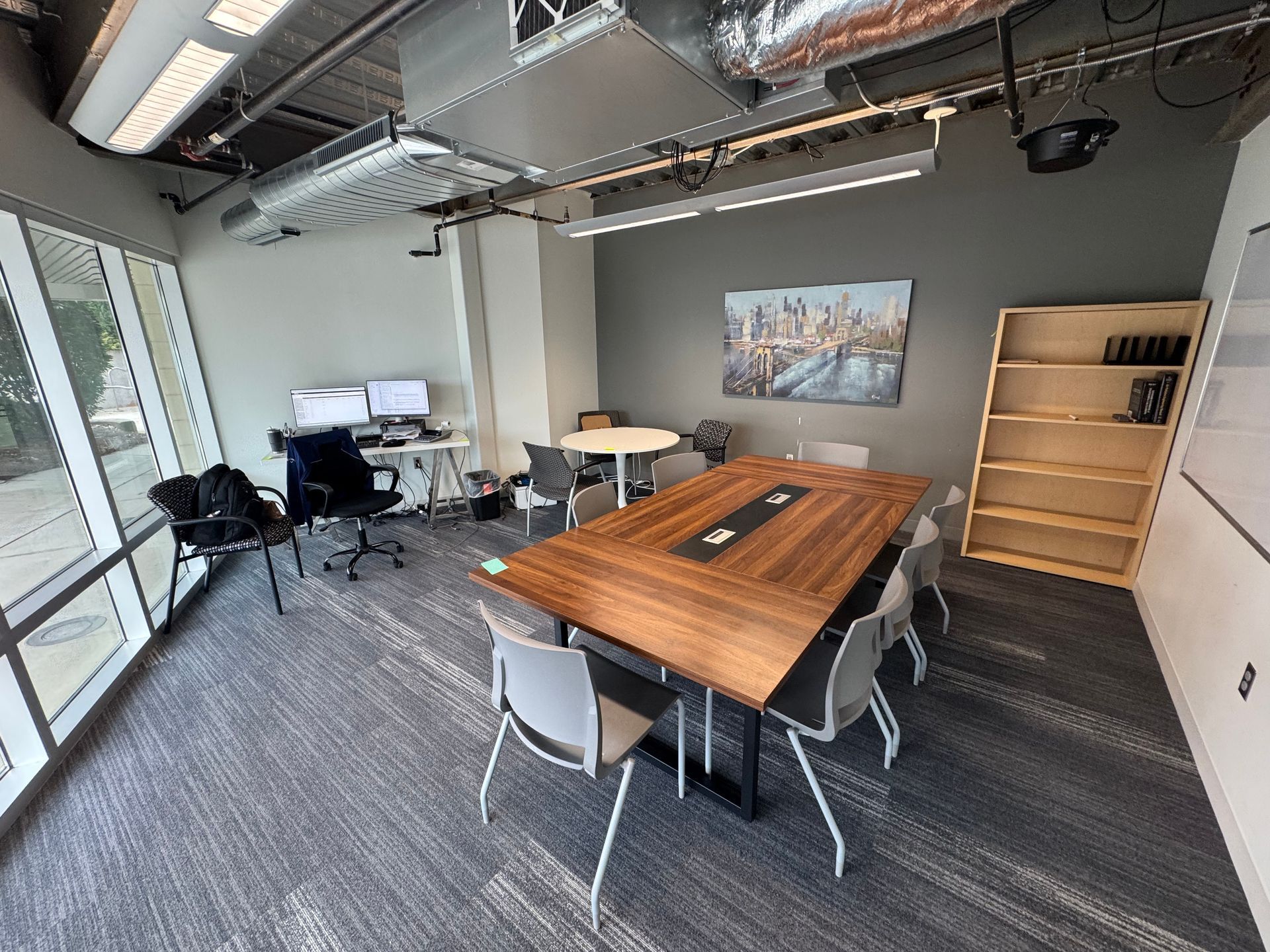 Conference room with long wooden table, gray chairs, white walls, and a bookshelf.