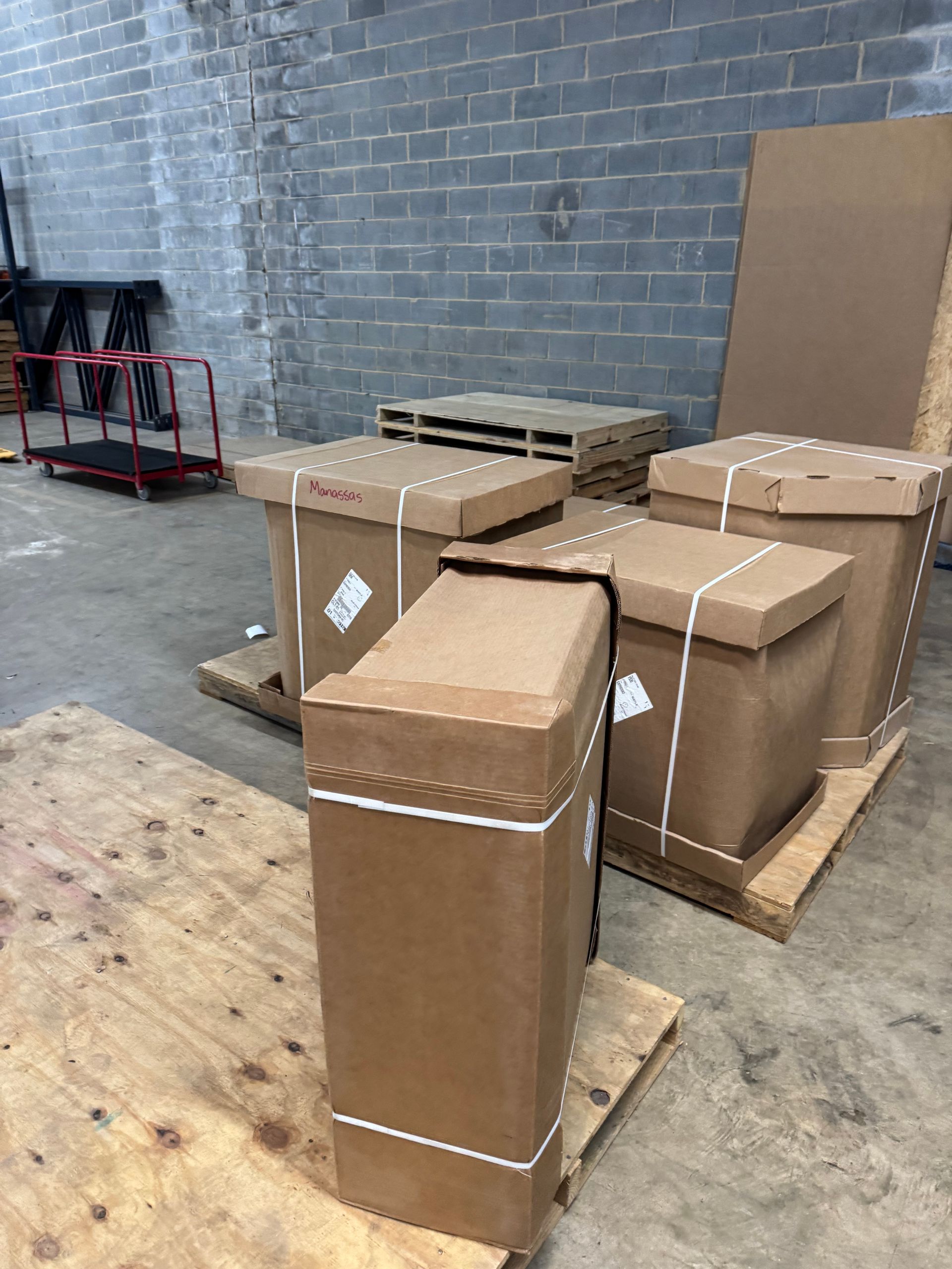 Cardboard boxes on pallets in a warehouse setting, secured with white straps.