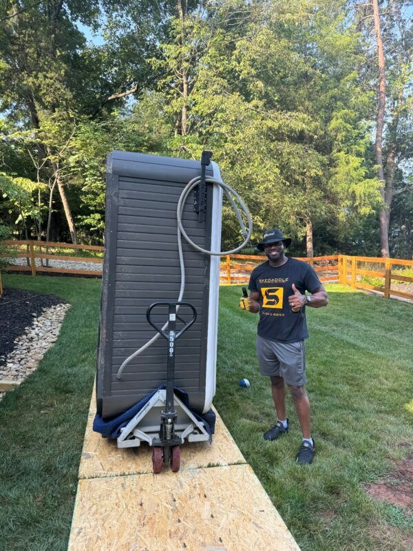 Man with thumbs up next to a hot tub on a pallet jack, outdoors on a sunny day.