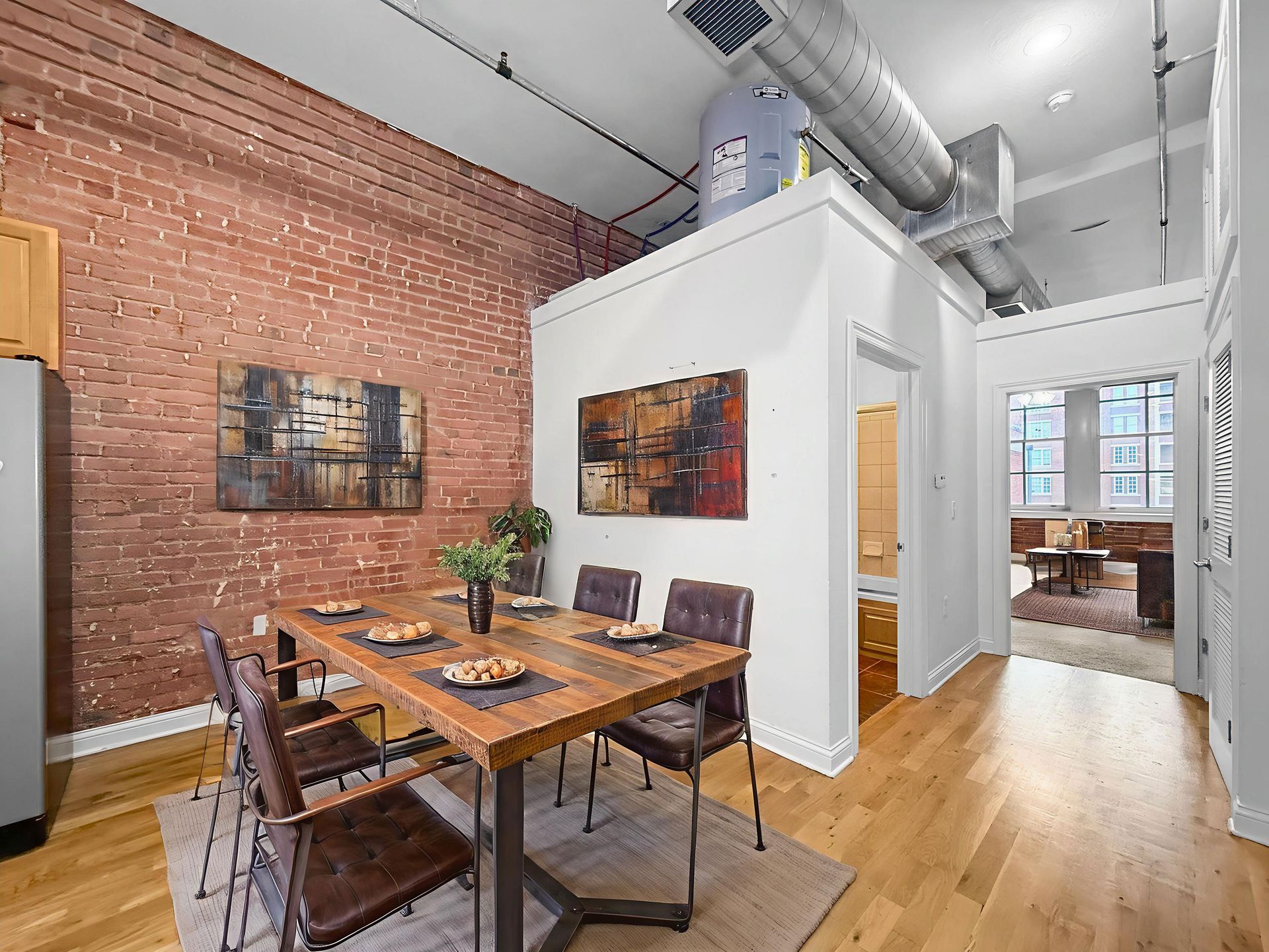 A wooden dining table with four brown chairs sits in a loft with exposed brick walls, white accents, and hardwood floors.