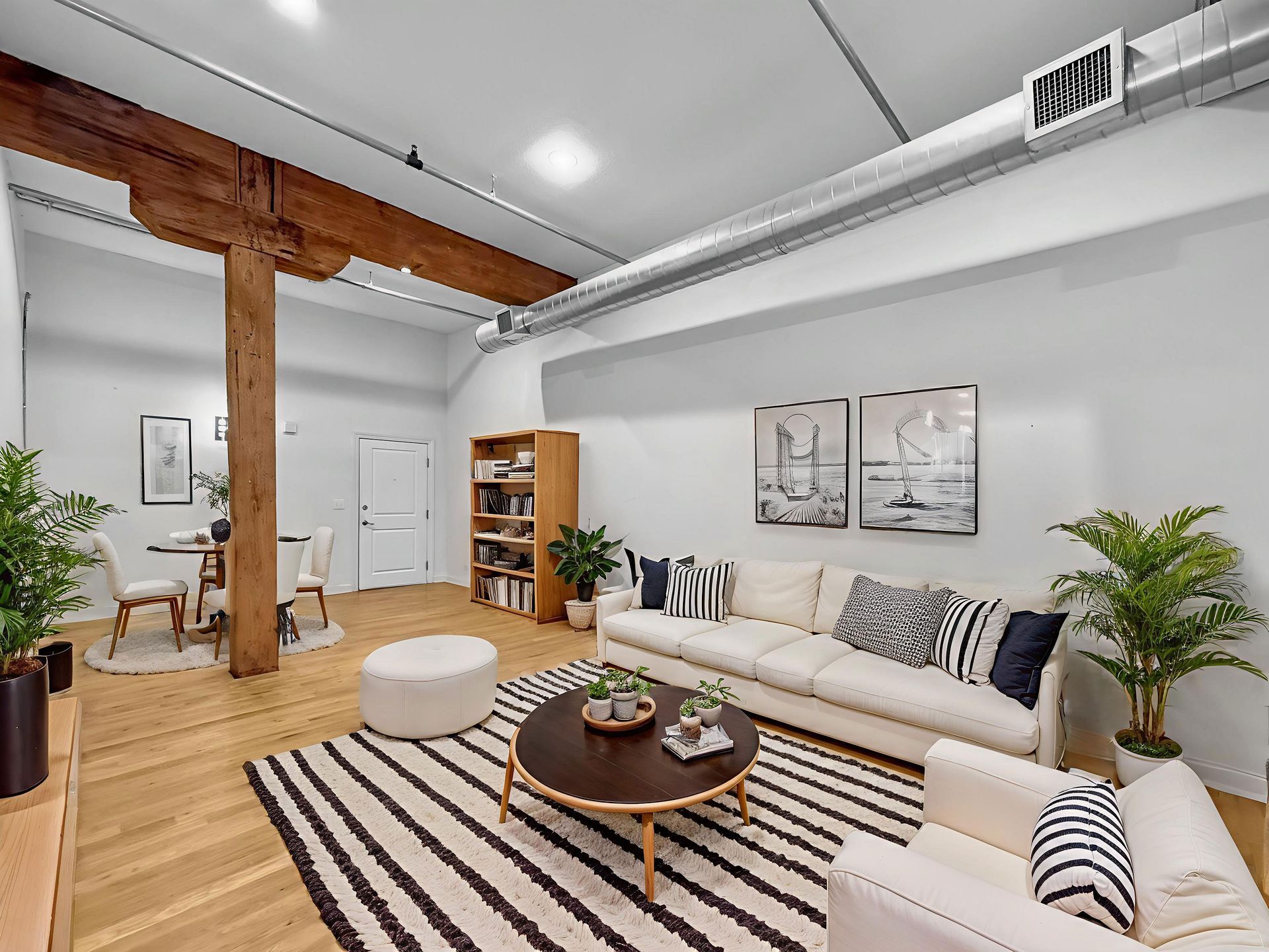 A bright, modern loft living room with a white sofa, round coffee table on a striped rug, wooden beam, and potted plants.
