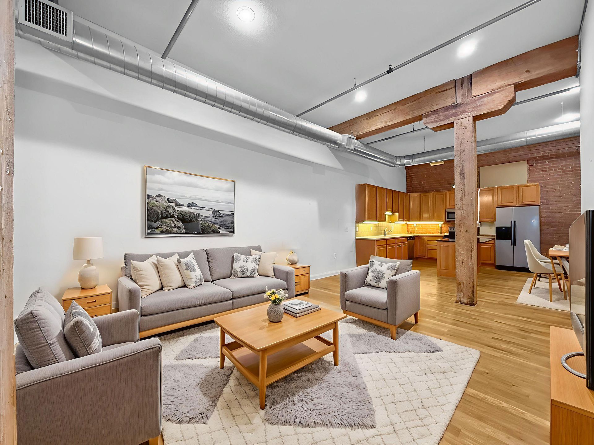 A modern open-concept living area with a grey sofa, matching chairs, wooden coffee table, and an exposed brick kitchen.