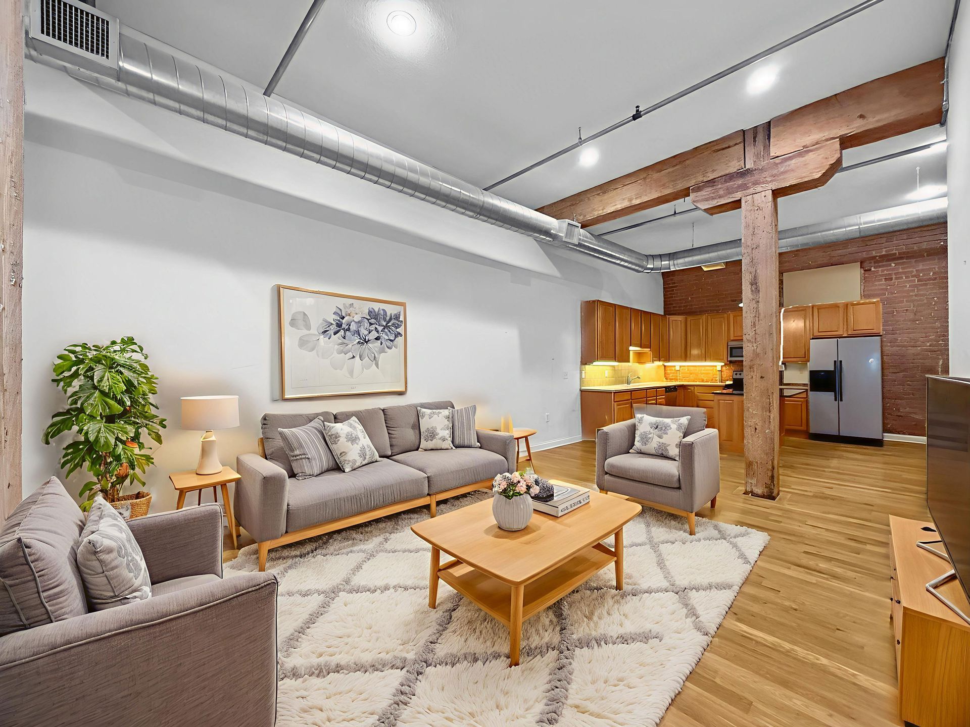 Modern loft living room with a grey sofa, armchair, coffee table, and area rug, featuring exposed brick and wooden beams.