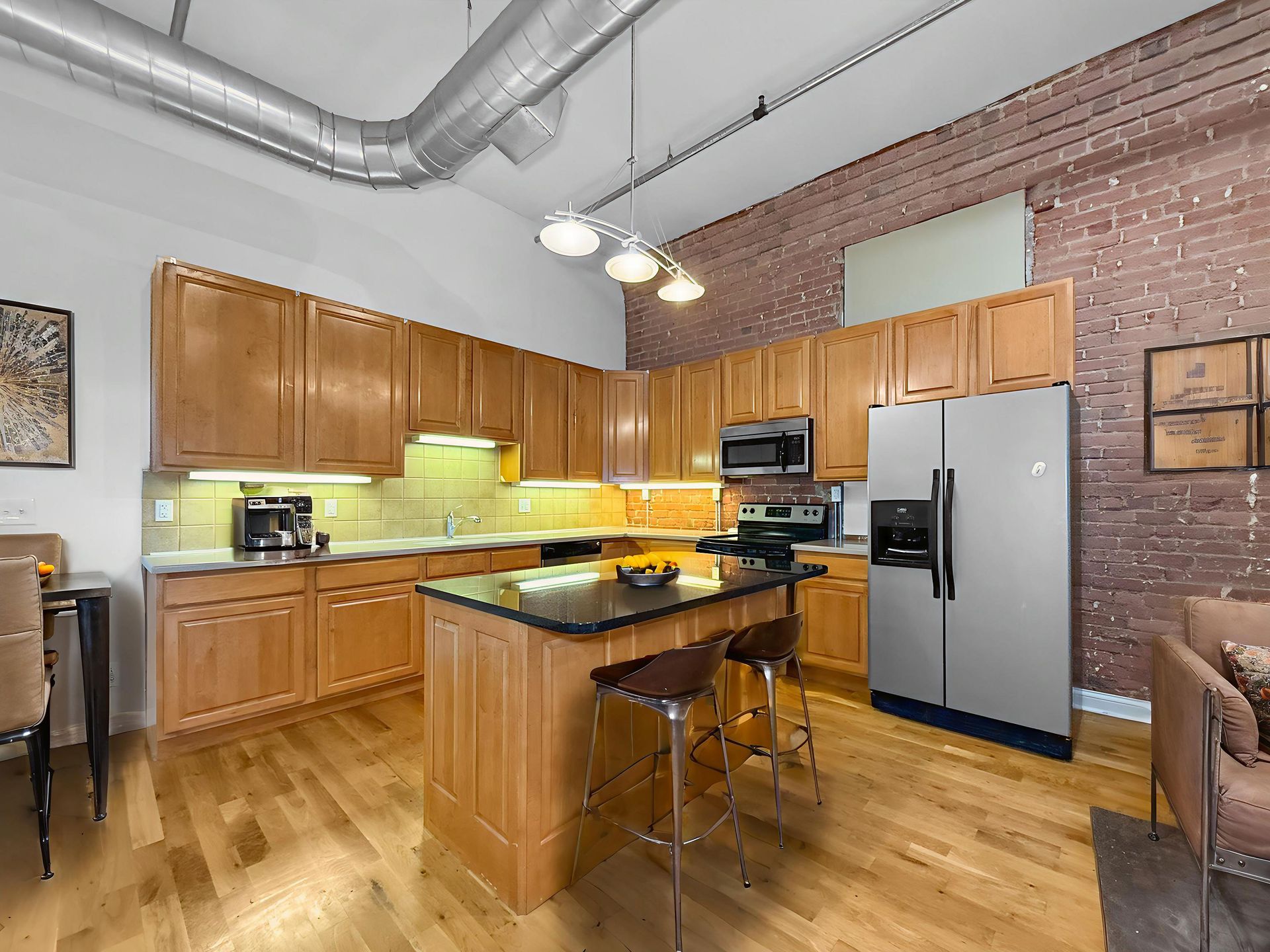 Modern kitchen featuring wooden cabinets, a dark island with bar stools, stainless steel appliances, and a brick wall.