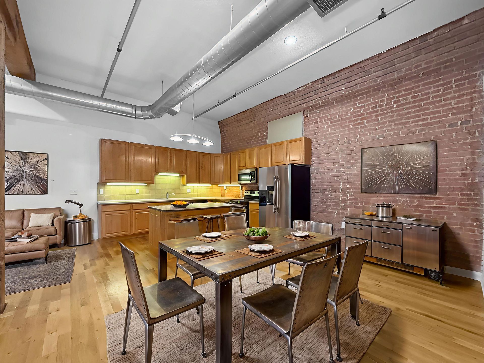 A modern open-plan apartment featuring a kitchen with wooden cabinets, a dining table, and an exposed brick wall.