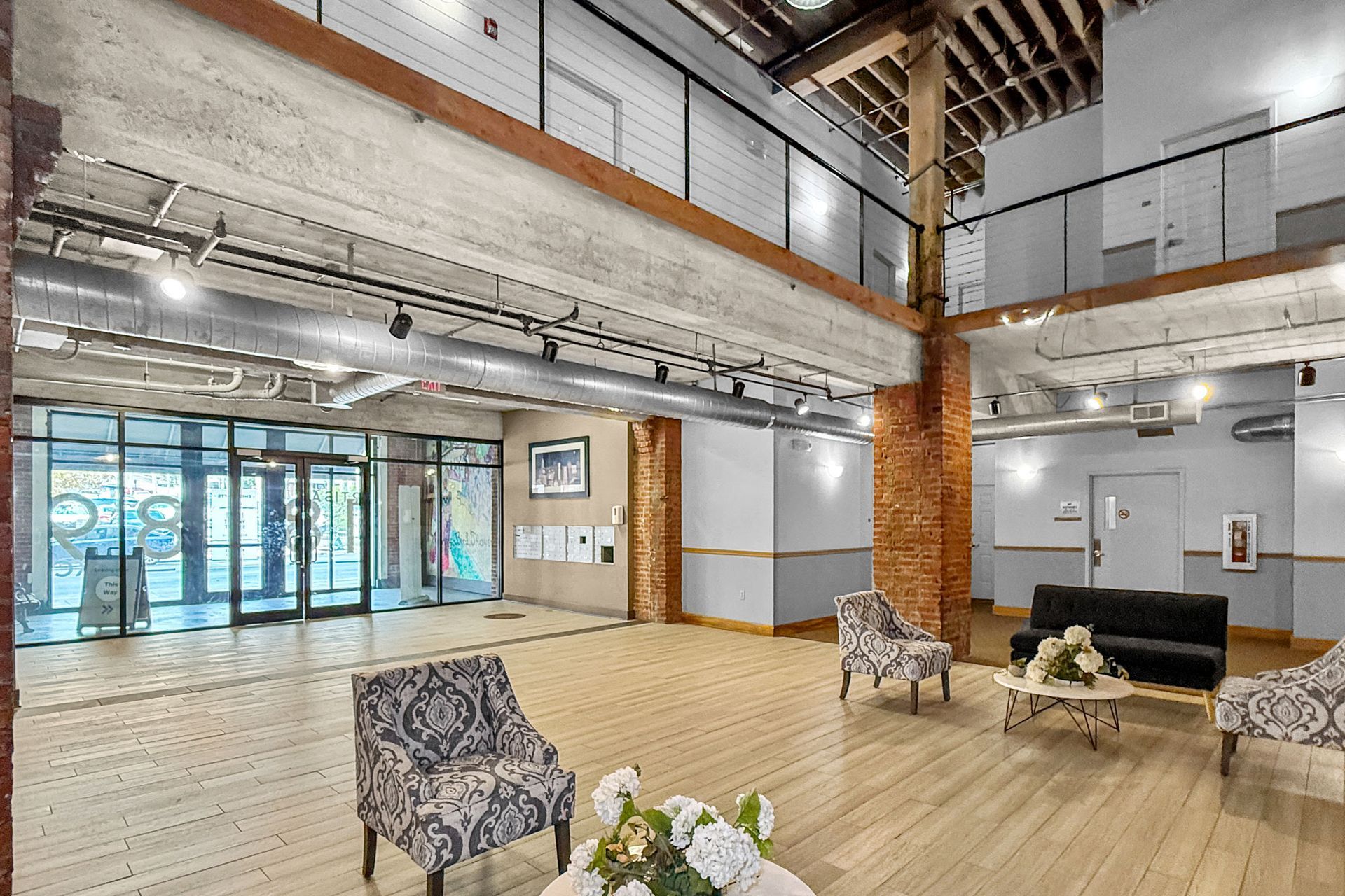 A spacious lobby with light wood floors, seating areas, large glass doors, and a lofted balcony with industrial beams.