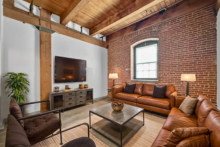 Living room with industrial design, featuring a large brick wall, exposed timber beams, leather sofas, and a TV console.