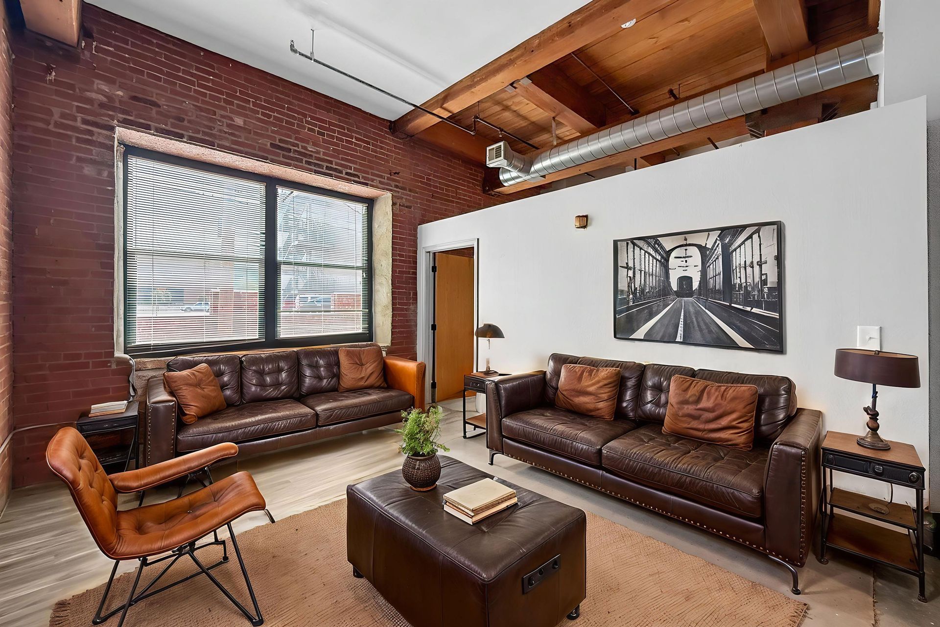 A loft living room with exposed brick walls, two brown leather sofas, a matching ottoman, and a framed wall print.