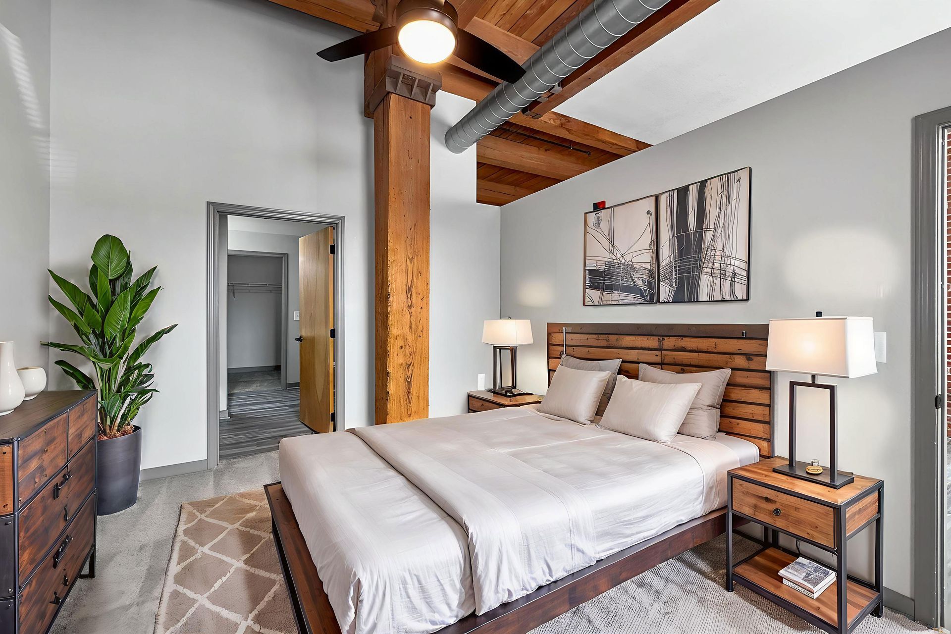 A modern bedroom with a wooden ceiling beam, a queen bed, matching nightstands, and a large plant in the corner.