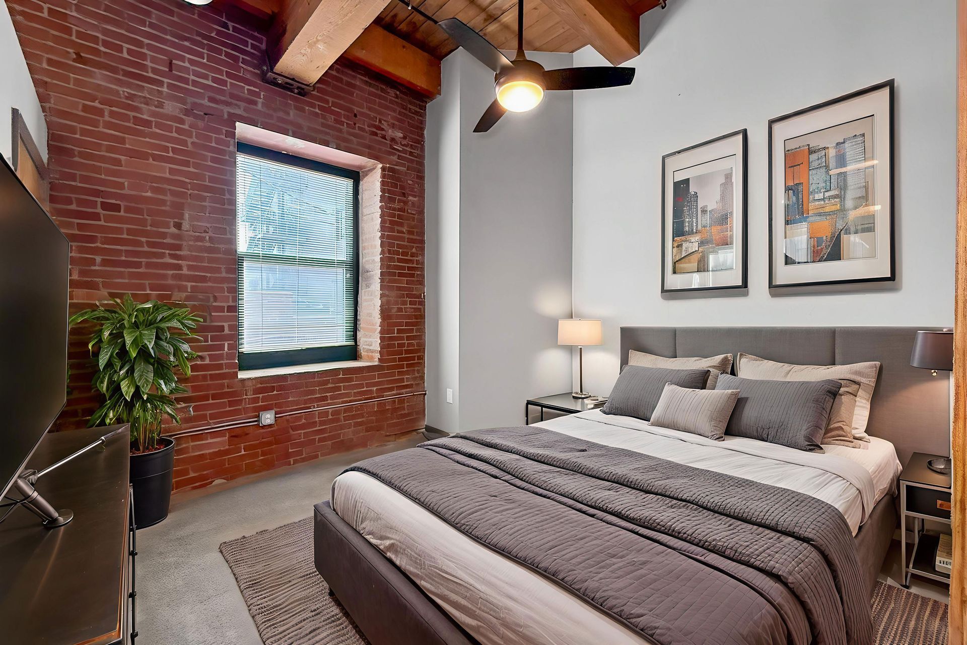 A bedroom with exposed brick walls, wooden ceiling beams, a bed with gray bedding, and two framed prints above the headboard.