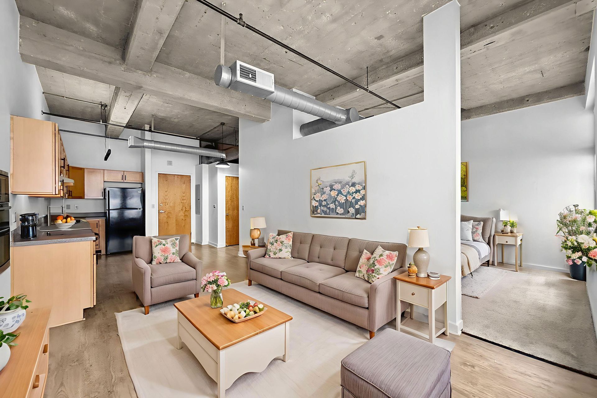 Modern loft living room with beige sofa, armchair, coffee table, and an adjacent open-concept kitchen and bedroom area.
