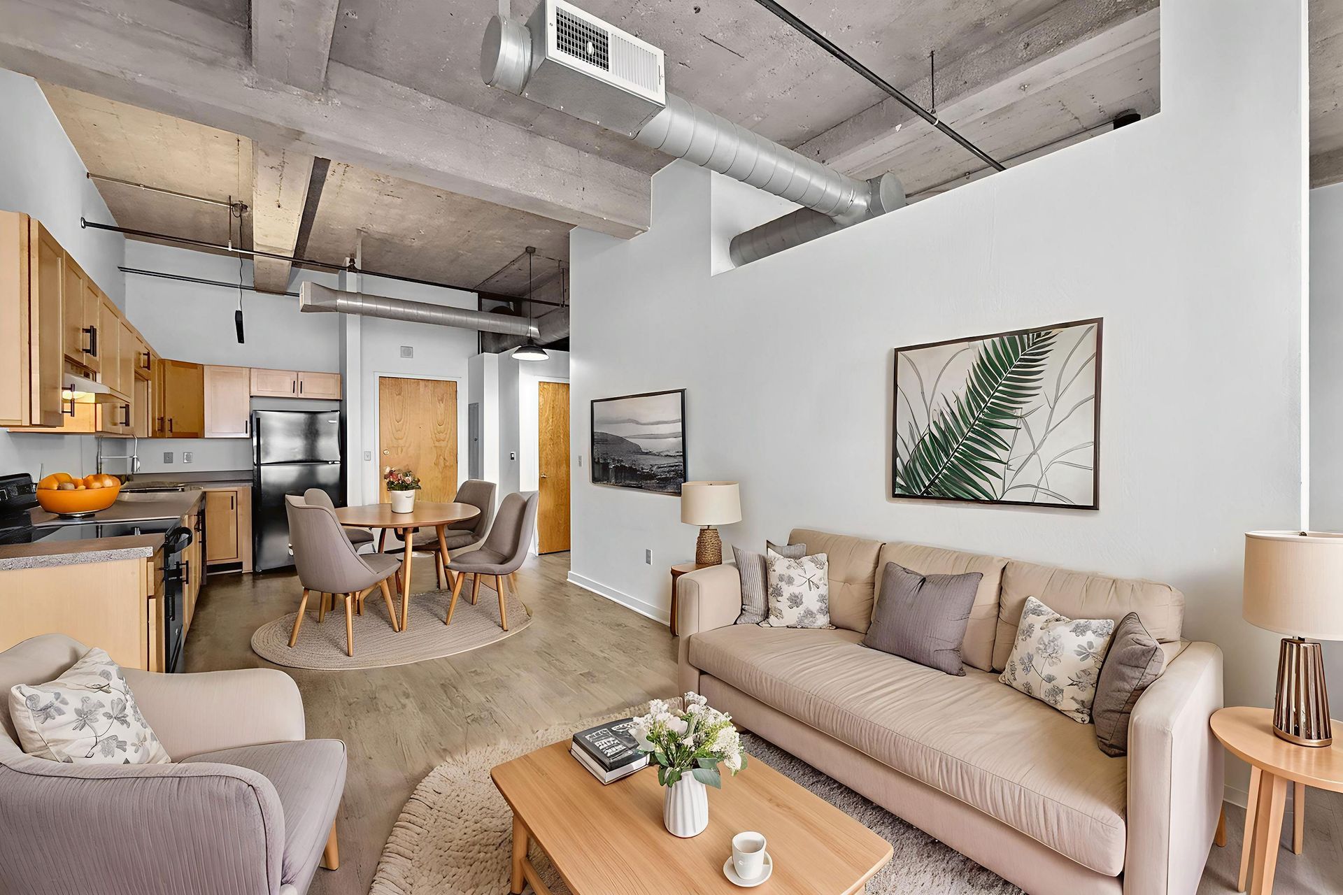 A modern open-plan apartment with a light-colored sofa, armchair, coffee table, dining set, and an industrial kitchen.